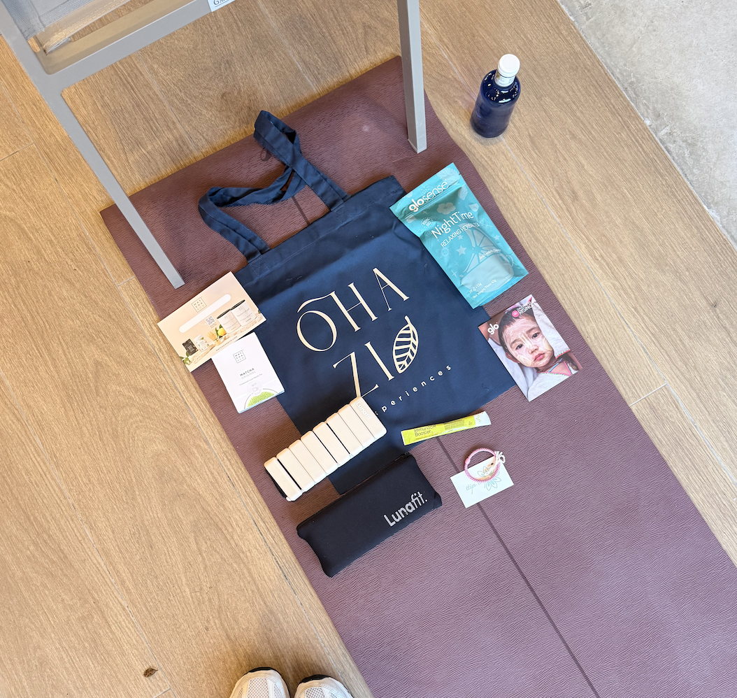 Varios objetos de bienestar y cuidado personal en una esterilla de yoga, incluyendo una bolsa de compras, una mascarilla nocturna, una crema, un paquete de vitaminas, un reloj, y una botella de agua, junto con unas zapatillas deportivas en la parte i