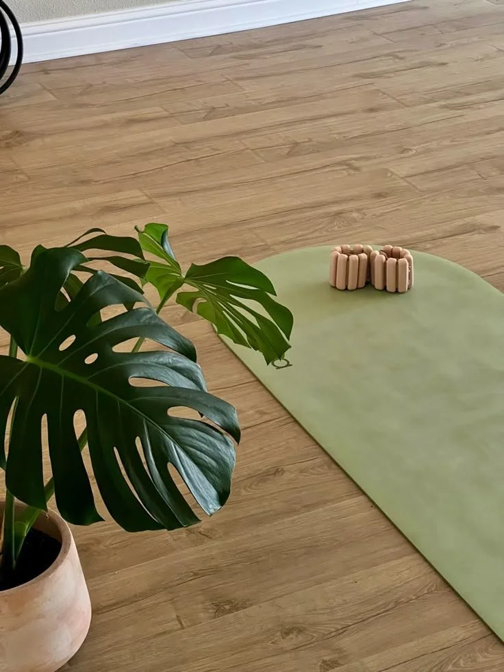 OHAZI EXPERIENCES: Un tapete de yoga verde en una habitación con piso de madera, con una pila de bloques de yoga de color beige y una planta grande de hojas verdes en una maceta de barro.