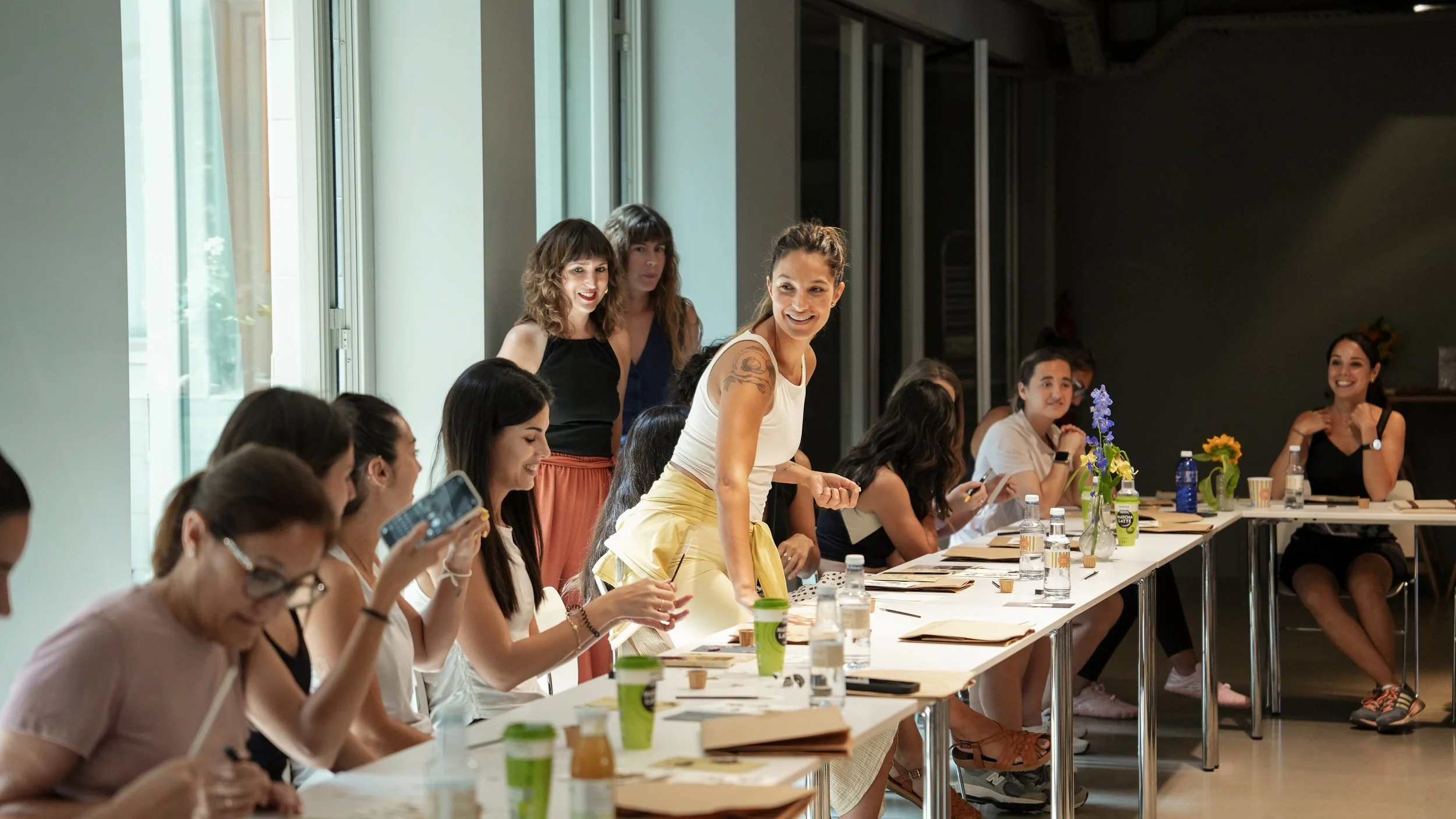 Grupo de mujeres en una reunión o taller, algunas conversando y otras usando teléfono en una mesa con flores y bebidas.