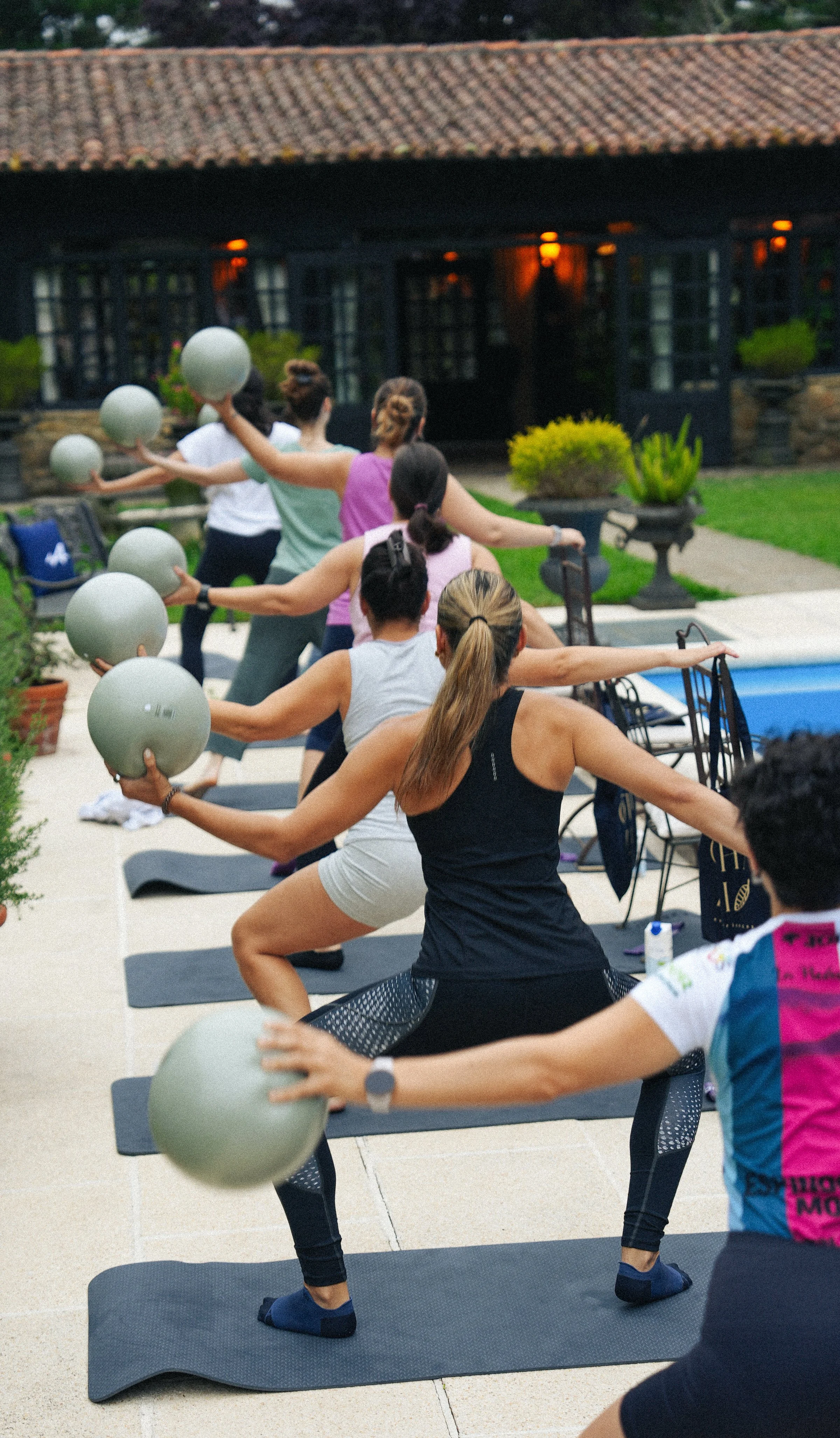 Grupo de personas haciendo yoga en exteriores con balones de ejercicio en las manos, en un patio con vegetación y un edificio de fondo.