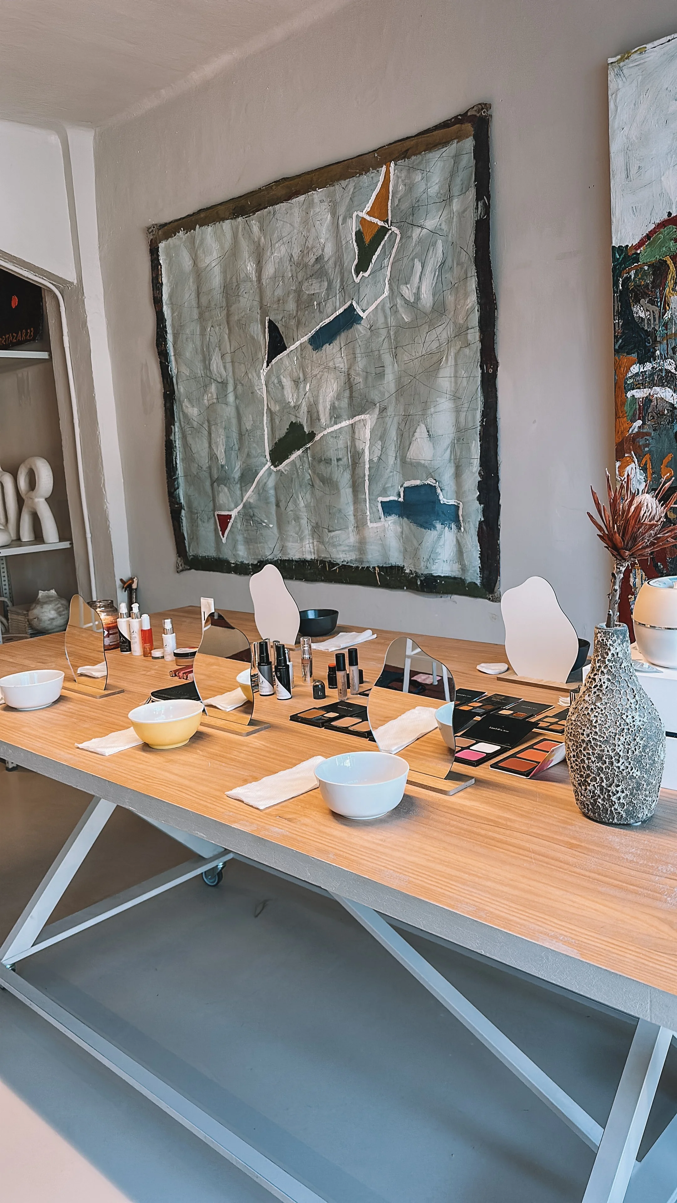 OHAZI EXPERIENCES: Mesa de maquillaje con espejos, productos de maquillaje y bowls blancos en una sala decorada con arte moderno y un jarrón con flores secas.