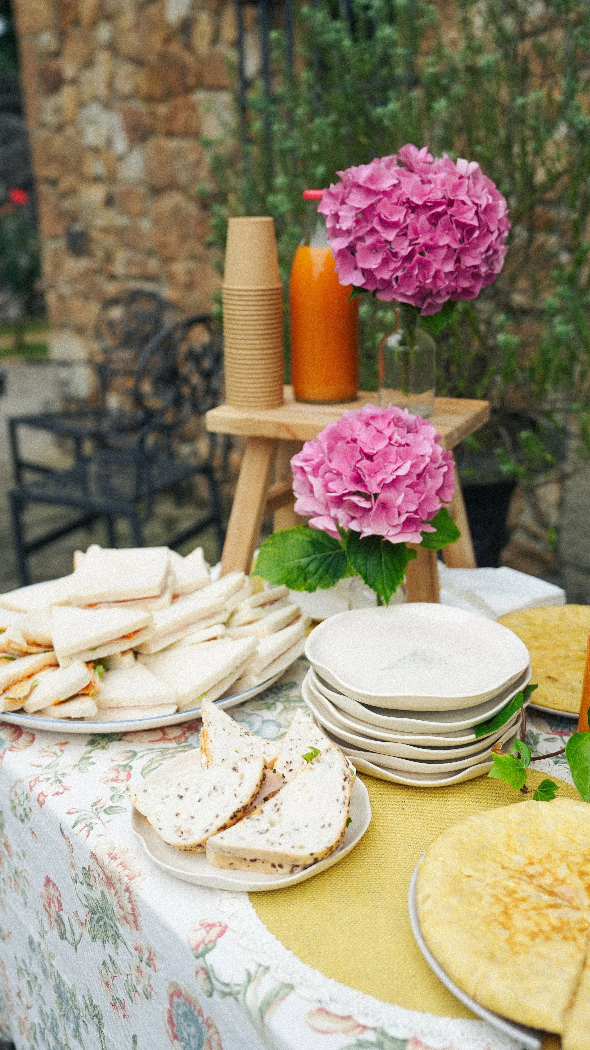 Mesa con sándwiches, pasteles y platos en un desayuno o comida, decorada con flores rosas y un fondo de jardín.