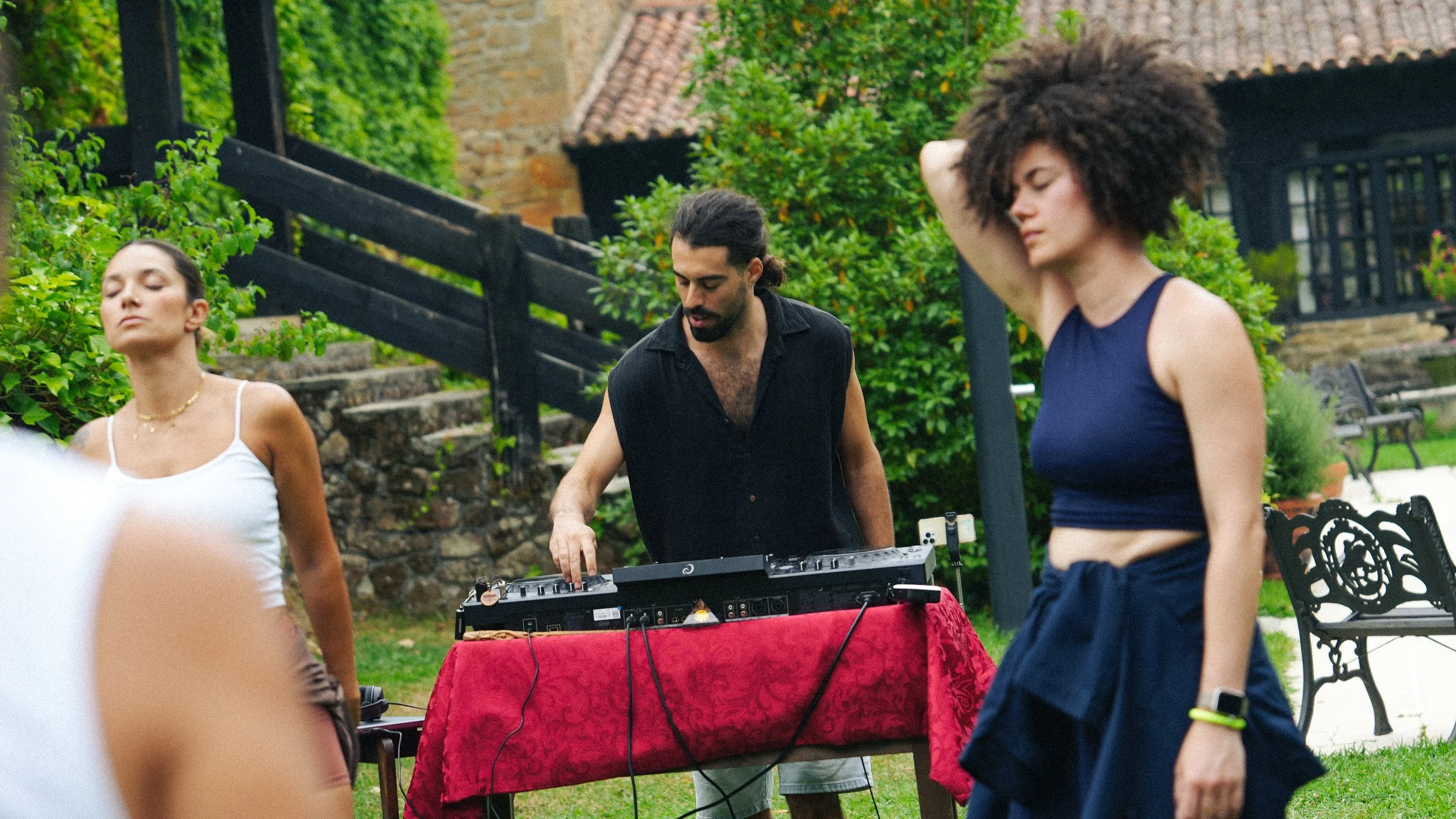 Tres personas bailando en un jardín, un hombre en medio ajustando equipo de música, mientras dos mujeres bailan con ojos cerrados, una con ropa blanca y otra con vestido azul, en un ambiente al aire libre con vegetación y escaleras de piedra.