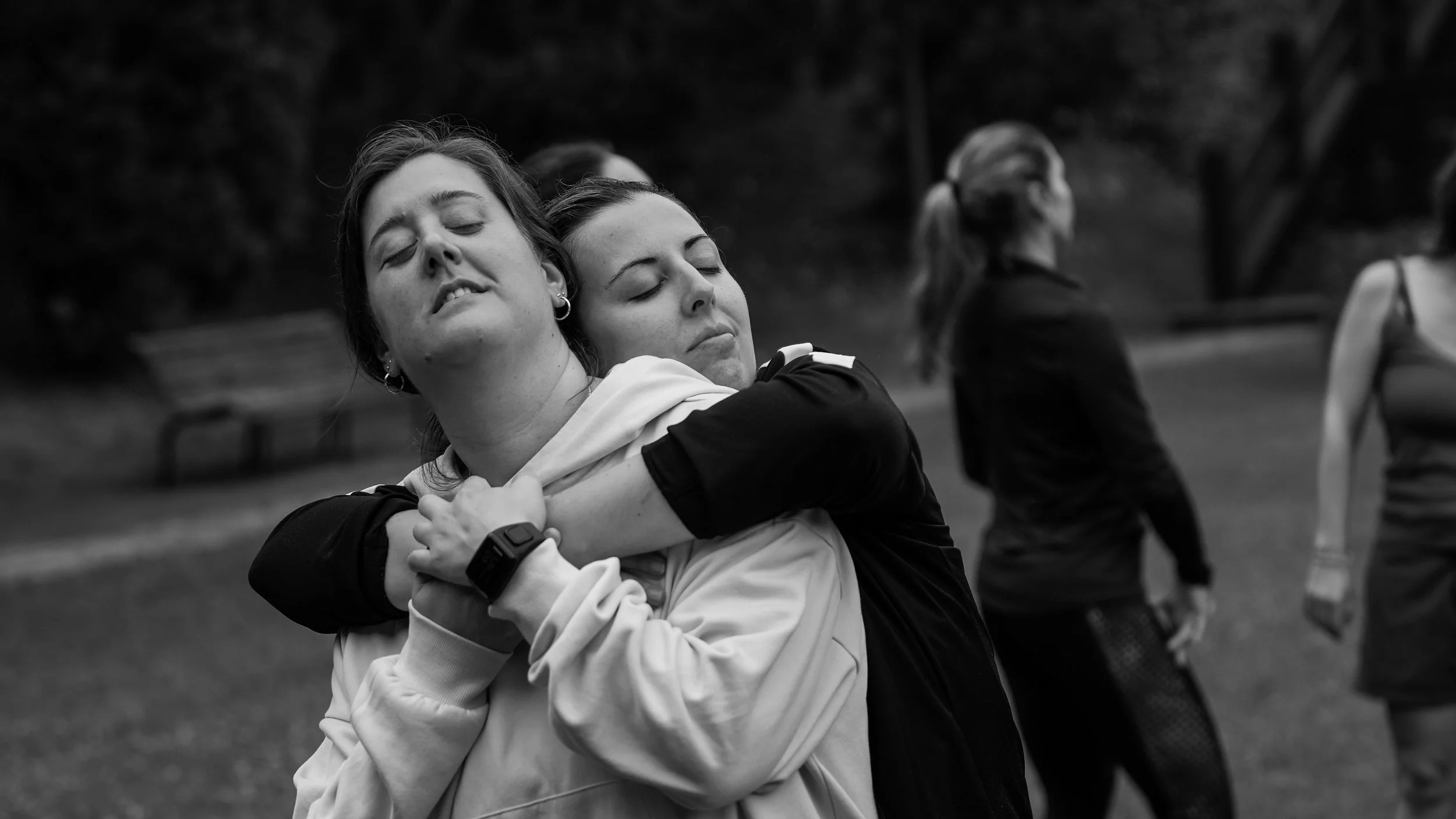 Dos mujeres abrazándose con los ojos cerrados, en un parque, en un entorno al aire libre.