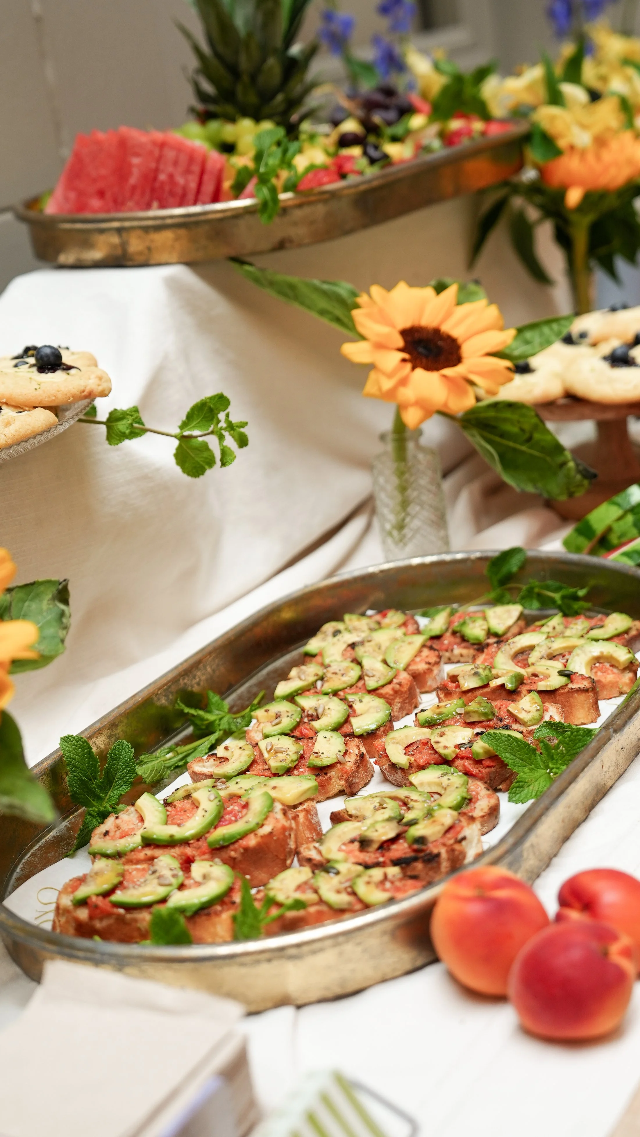 Mesa decorada con flores, frutas y alimentos como pan con aguacate y tomate, y galletas con arándanos.