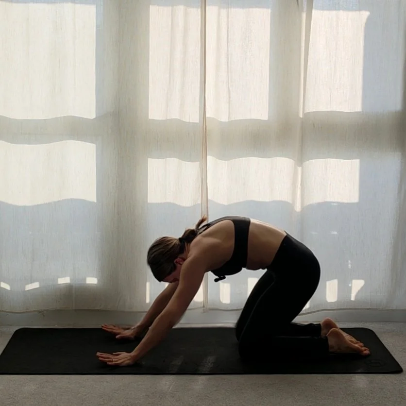 A woman practicing yoga, kneeling on a black mat in front of a white curtain, with her hands and knees on the mat, and her torso bent forward.