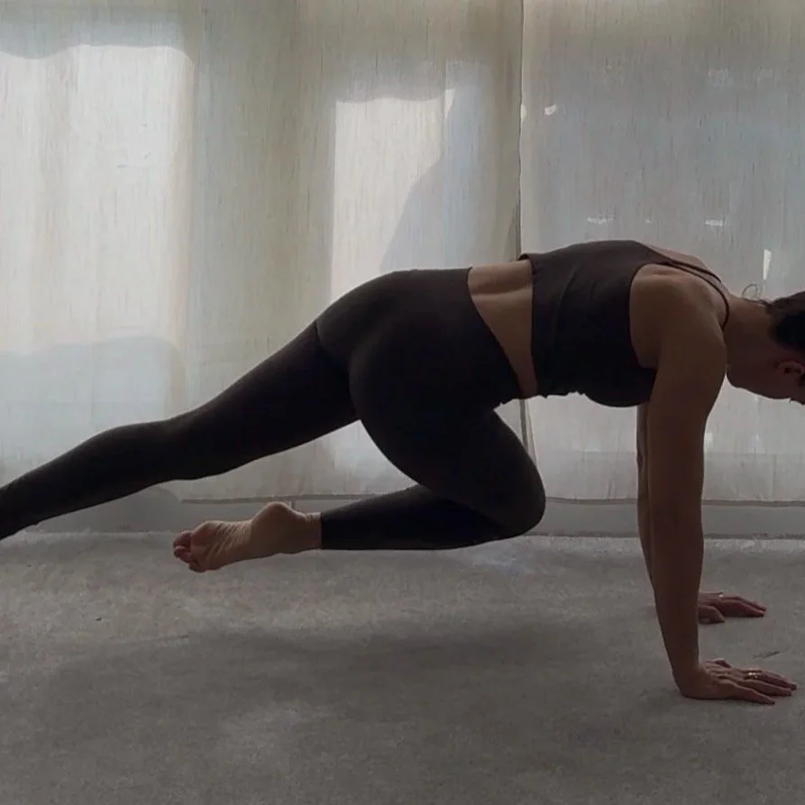 A woman practicing yoga indoors on a gray carpet in front of a sheer curtain, performing a plank pose with her knees off the ground and back straight.