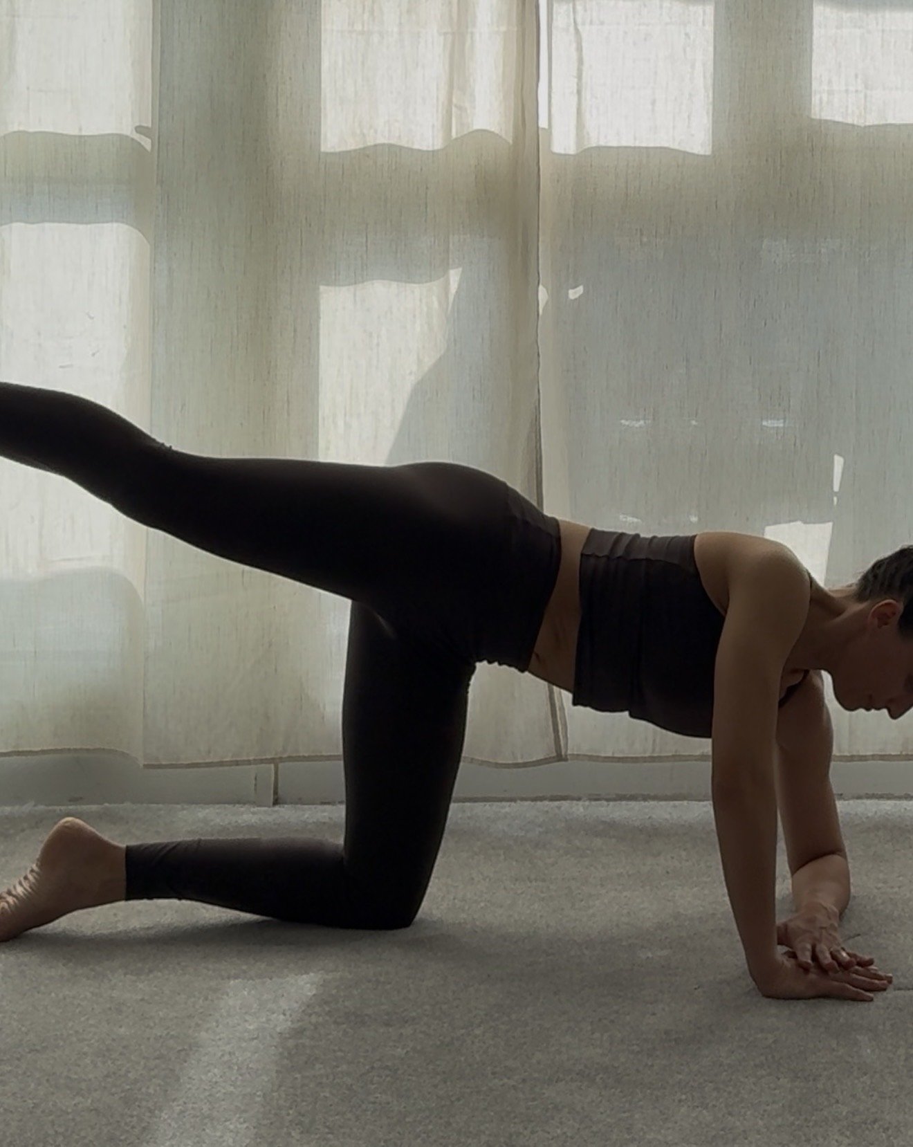 A person practicing yoga on a carpeted floor, in a tabletop position with one leg extended back. They are wearing black athletic clothing and are in a room with sheer curtains allowing natural light to fill the space.