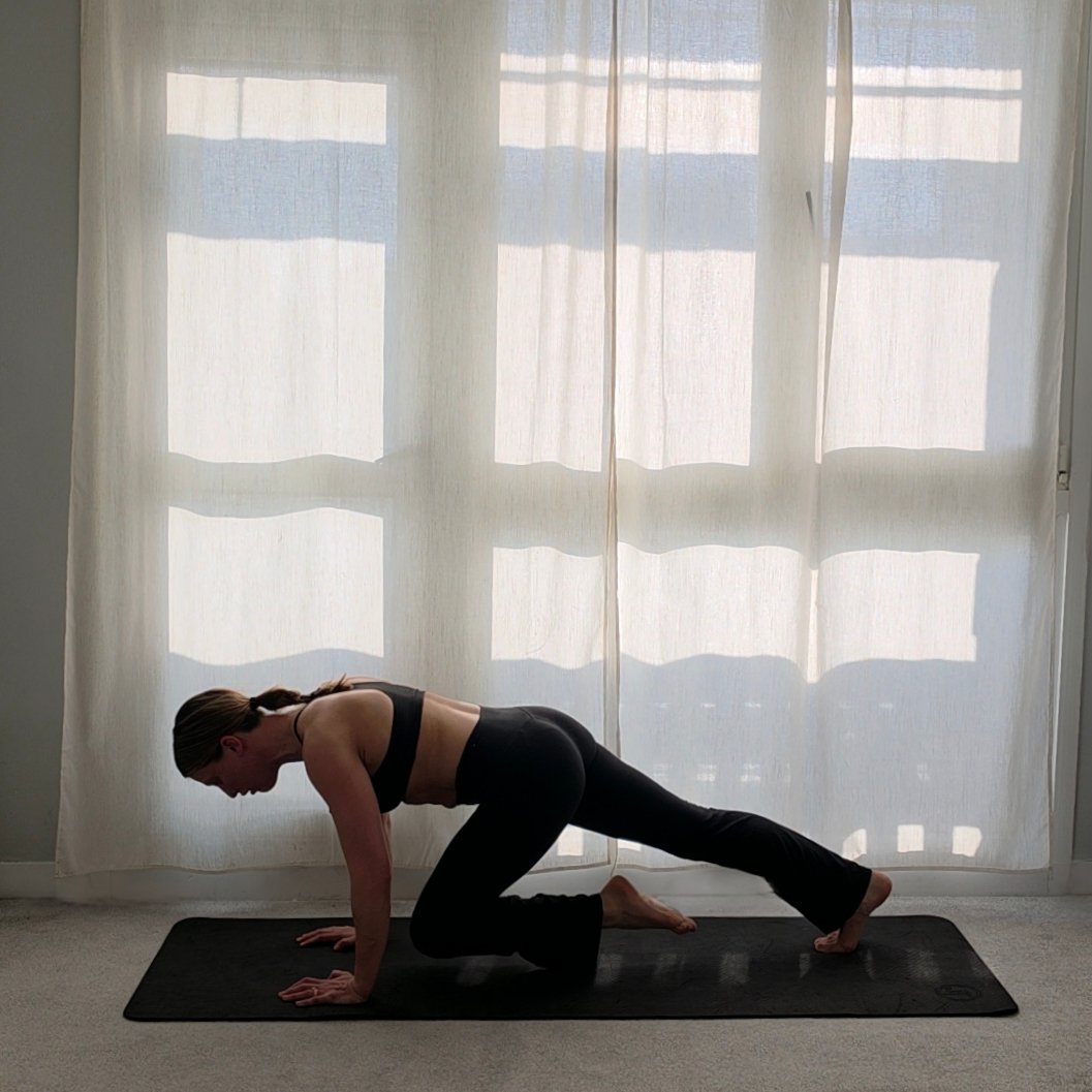 A woman in workout clothes practicing yoga or exercises on a black mat indoors in front of a sheer white curtain.