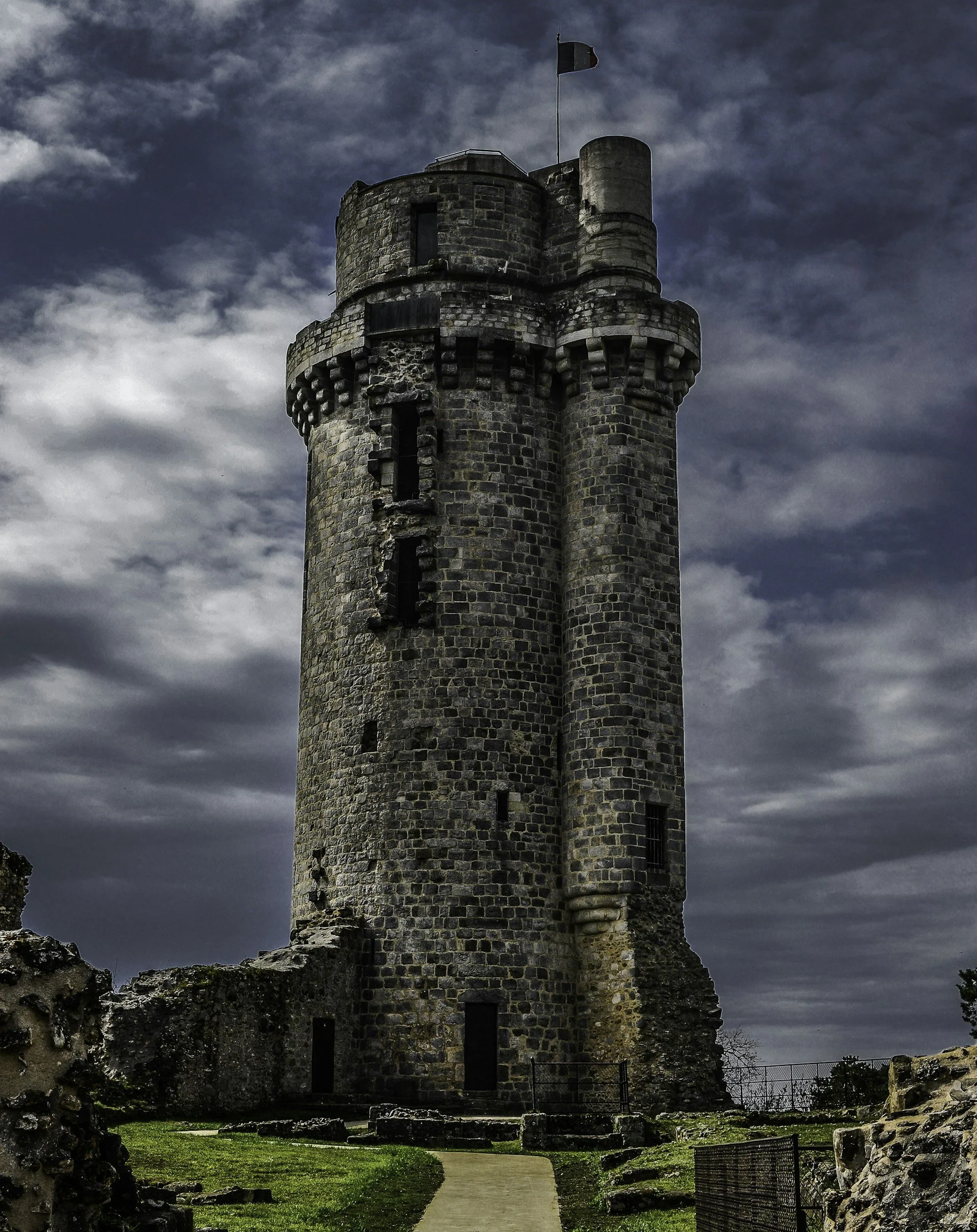 A tall, medieval stone tower with a flag on top, set against a cloudy sky. The tower has small windows and a walkway around its upper level.