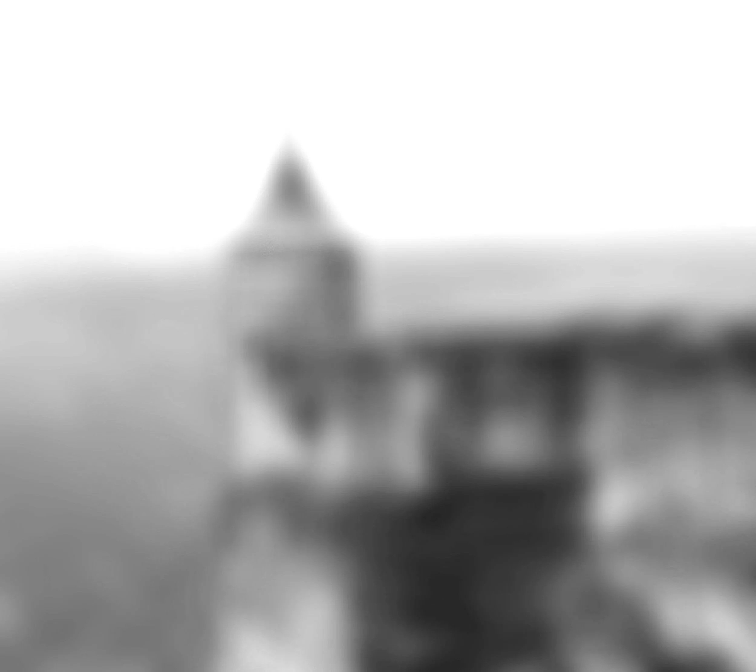 Blurred black and white photo of a building with a pointed roof, reflected in calm water.