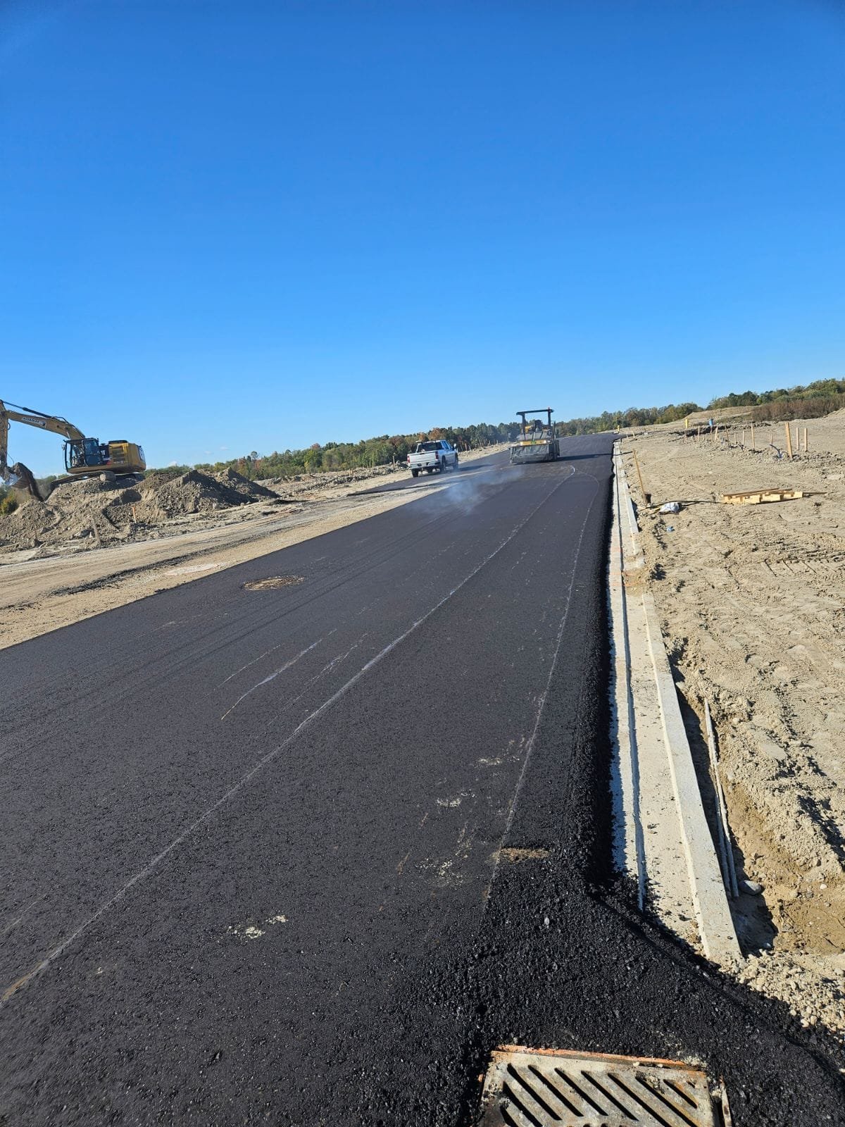 Newly paved black asphalt road under construction with a construction vehicle and a worker roller on the road, dirt piles on sides, and a pickup truck in the distance on a clear day.