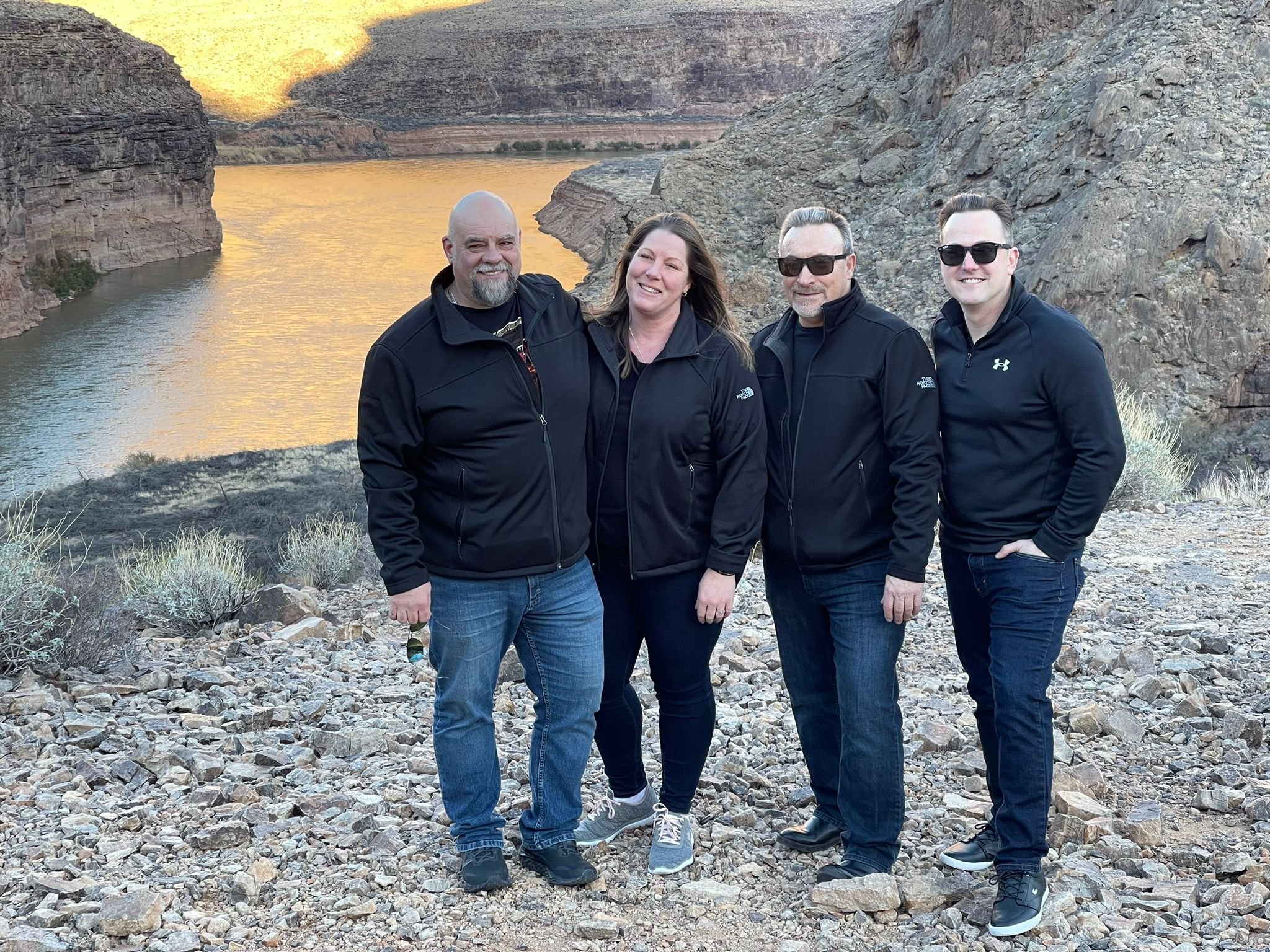 Four people standing together on rocky terrain with a river and canyon behind them during sunset, all wearing black jackets and sunglasses.