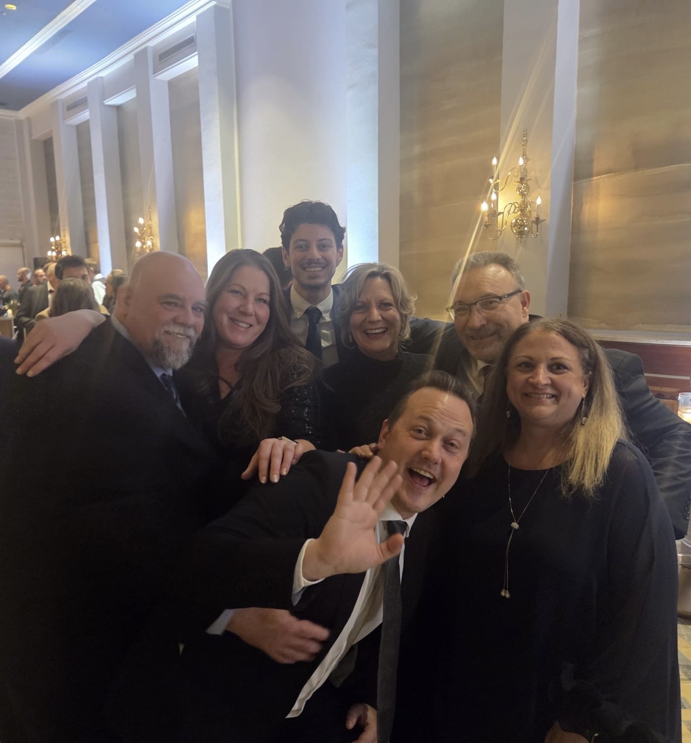 Group of seven people smiling and posing together at a formal event in an elegant room with chandeliers and tall decor.