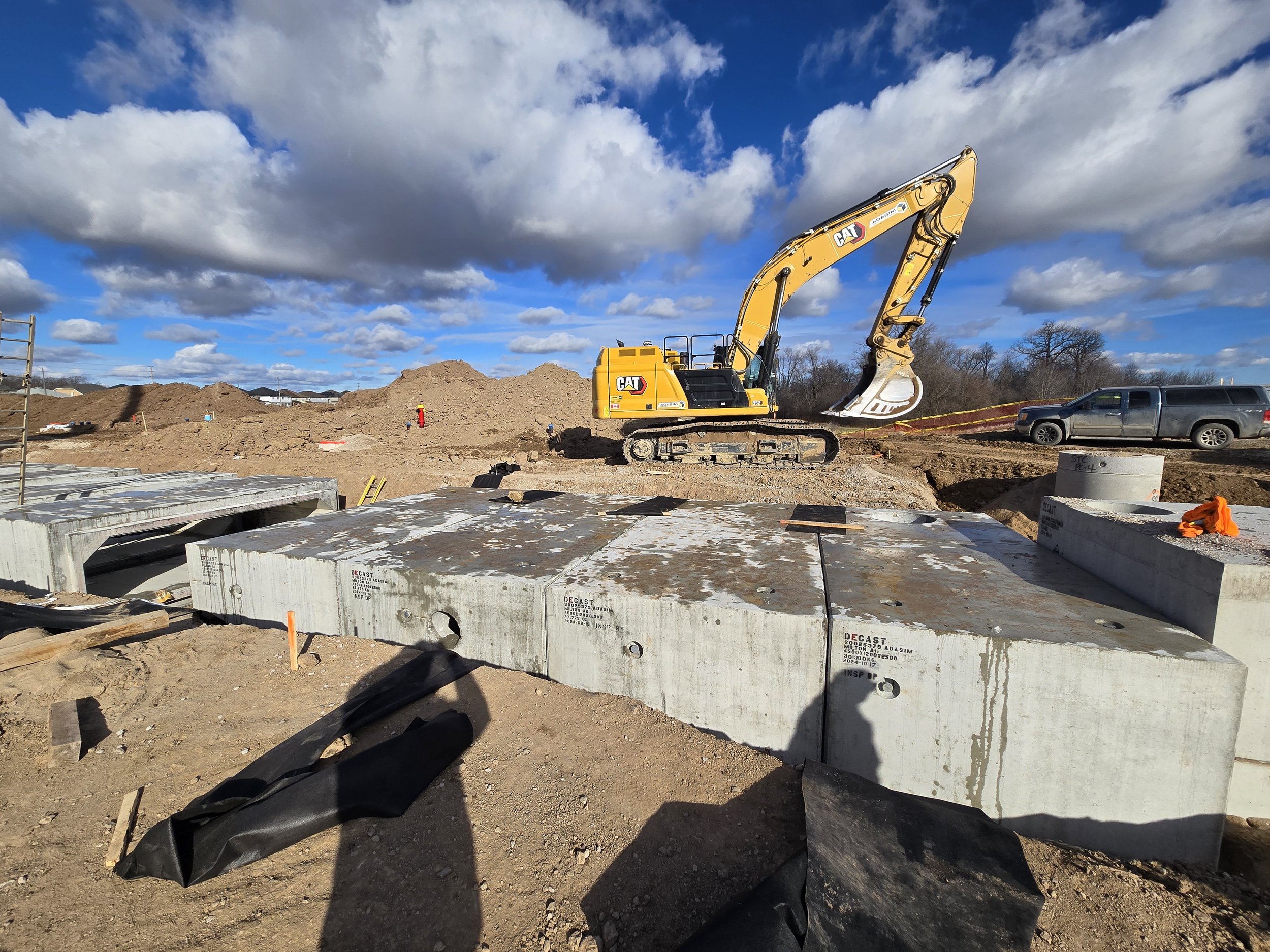 Construction site with a yellow Caterpillar excavator, large concrete blocks, dirt piles, a pickup truck, and a cloudy blue sky.