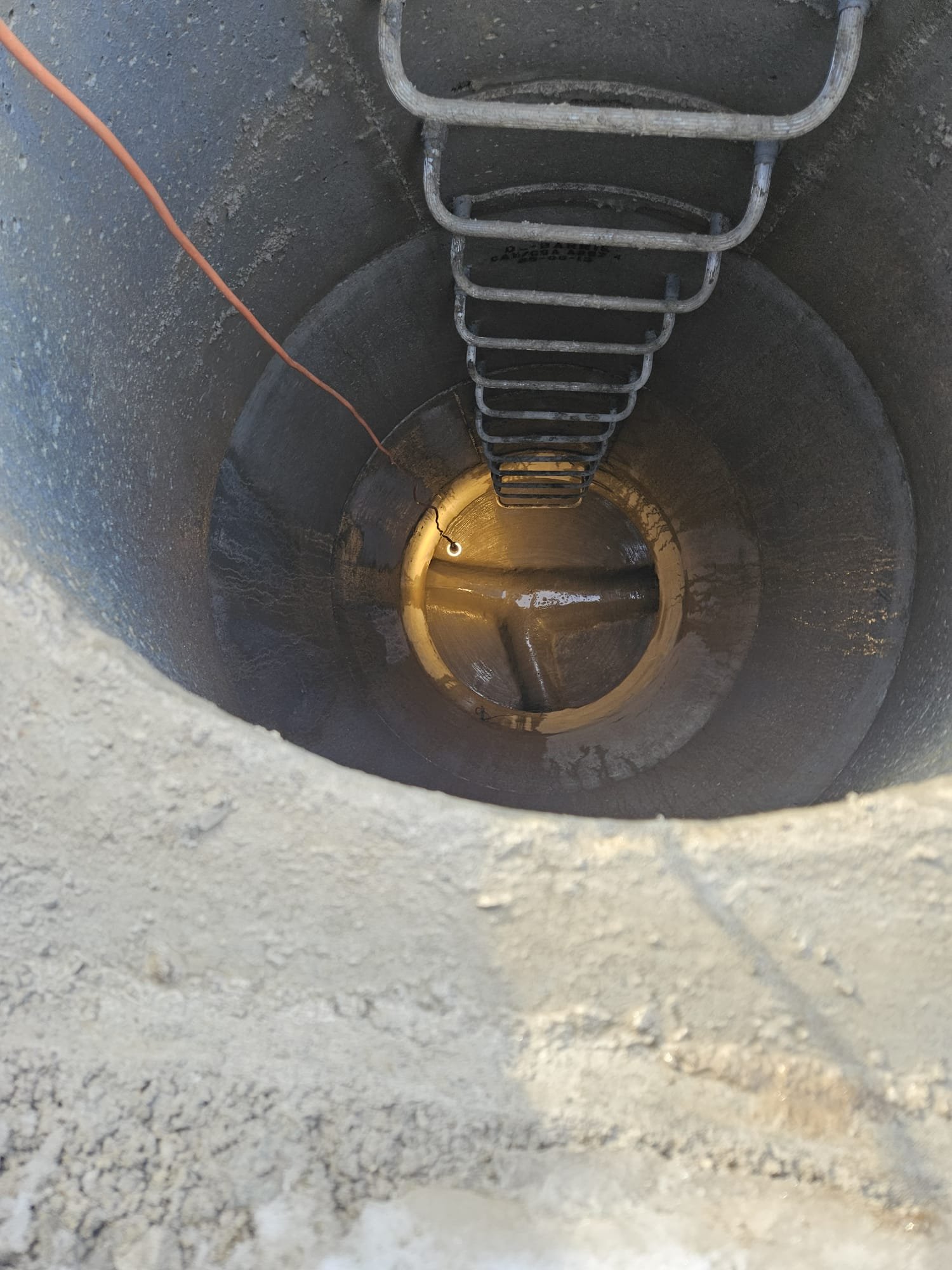 View down a deep, round well with a metal ladder attached to the side for climbing out. The sides are concrete and the bottom is wet, with some water pooled. An orange wire or cable is hanging into the well from above.