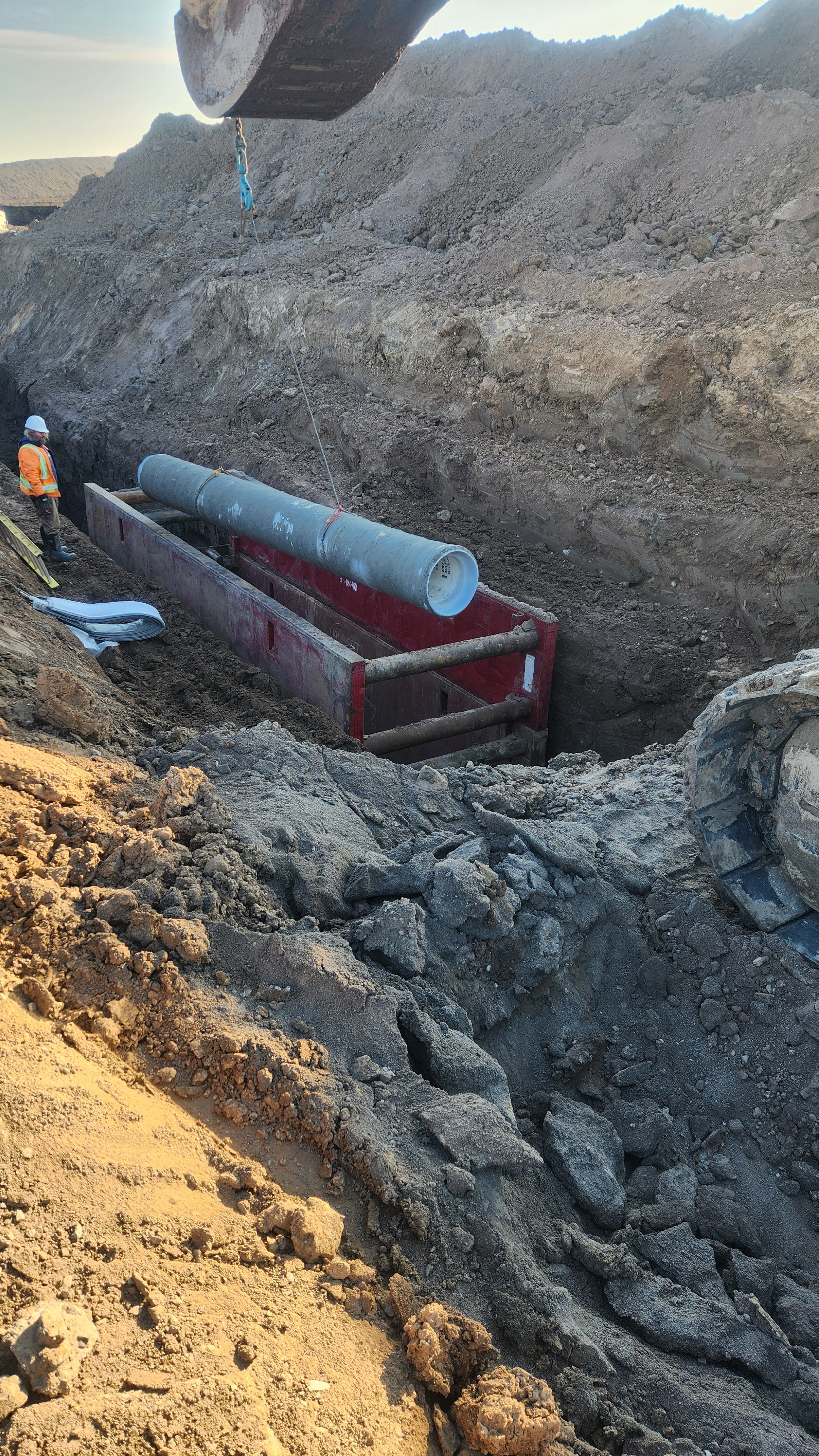 Construction workers install a large pipe into a trench at a construction site, with heavy equipment and dirt piles surrounding them.