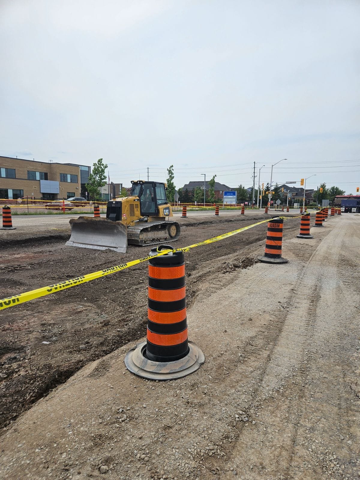 Construction site on a road with orange and black traffic barrels, yellow caution tape, and a small bulldozer, with buildings and traffic signals in the background.