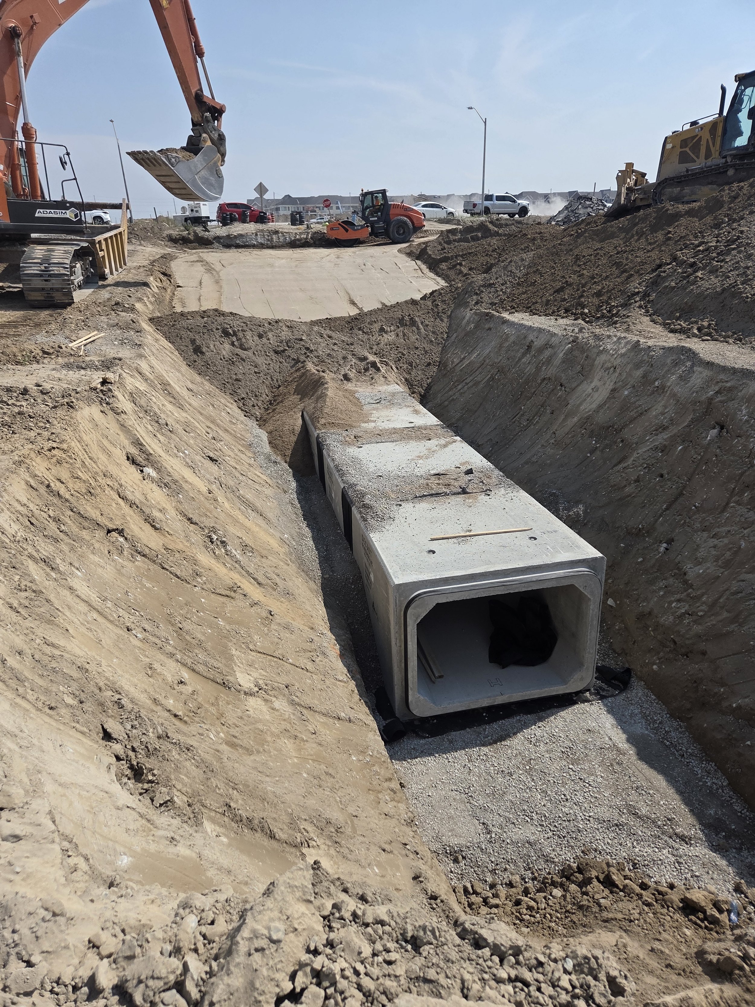 Construction site with heavy machinery, including an excavator and a roller, working on a dirt road with a large concrete pipe placed in a trench.