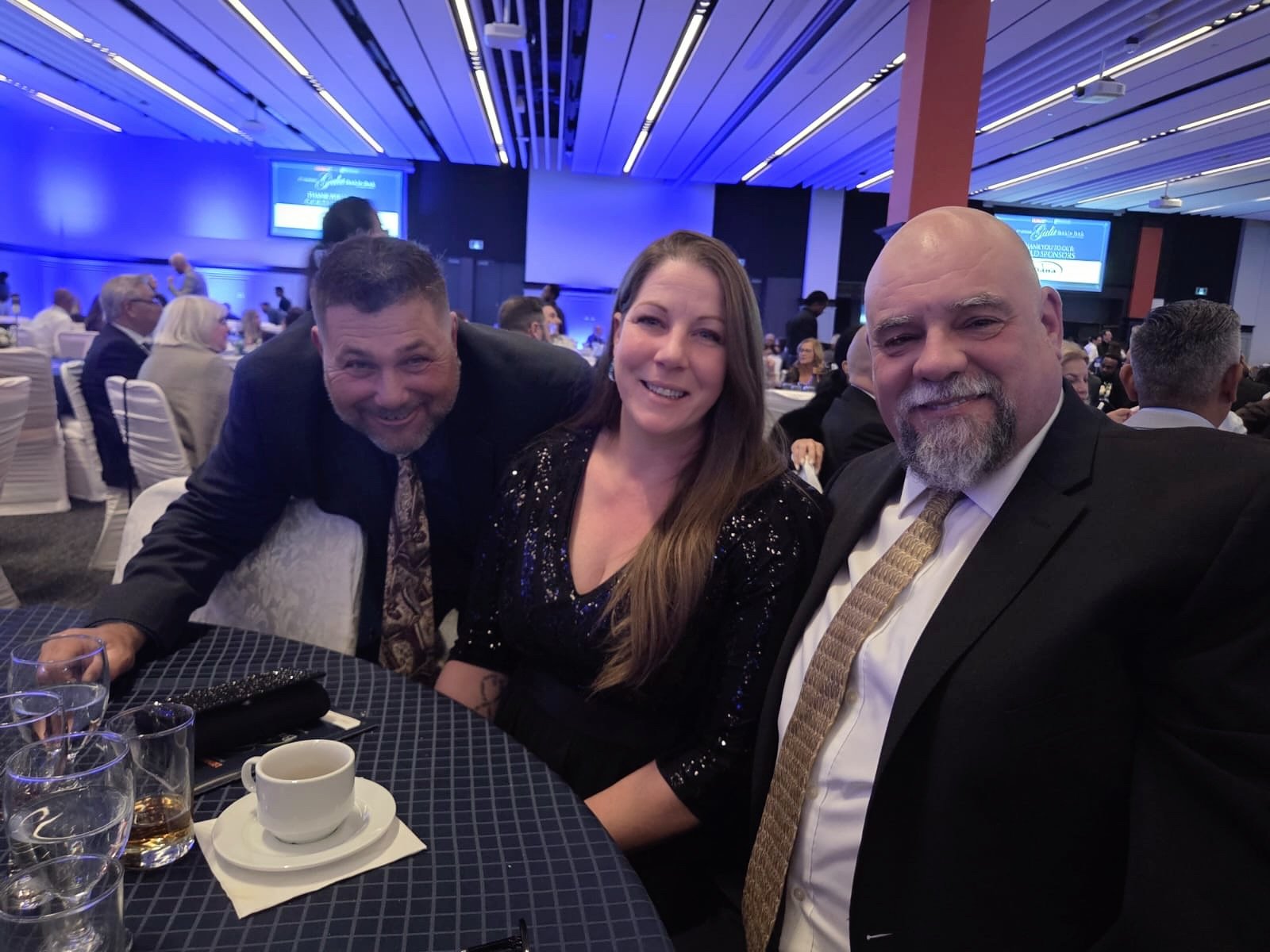 Three people, two men and one woman, dressed in formal attire, sitting together at a round table in a large banquet hall with blue lighting and many other guests in the background.