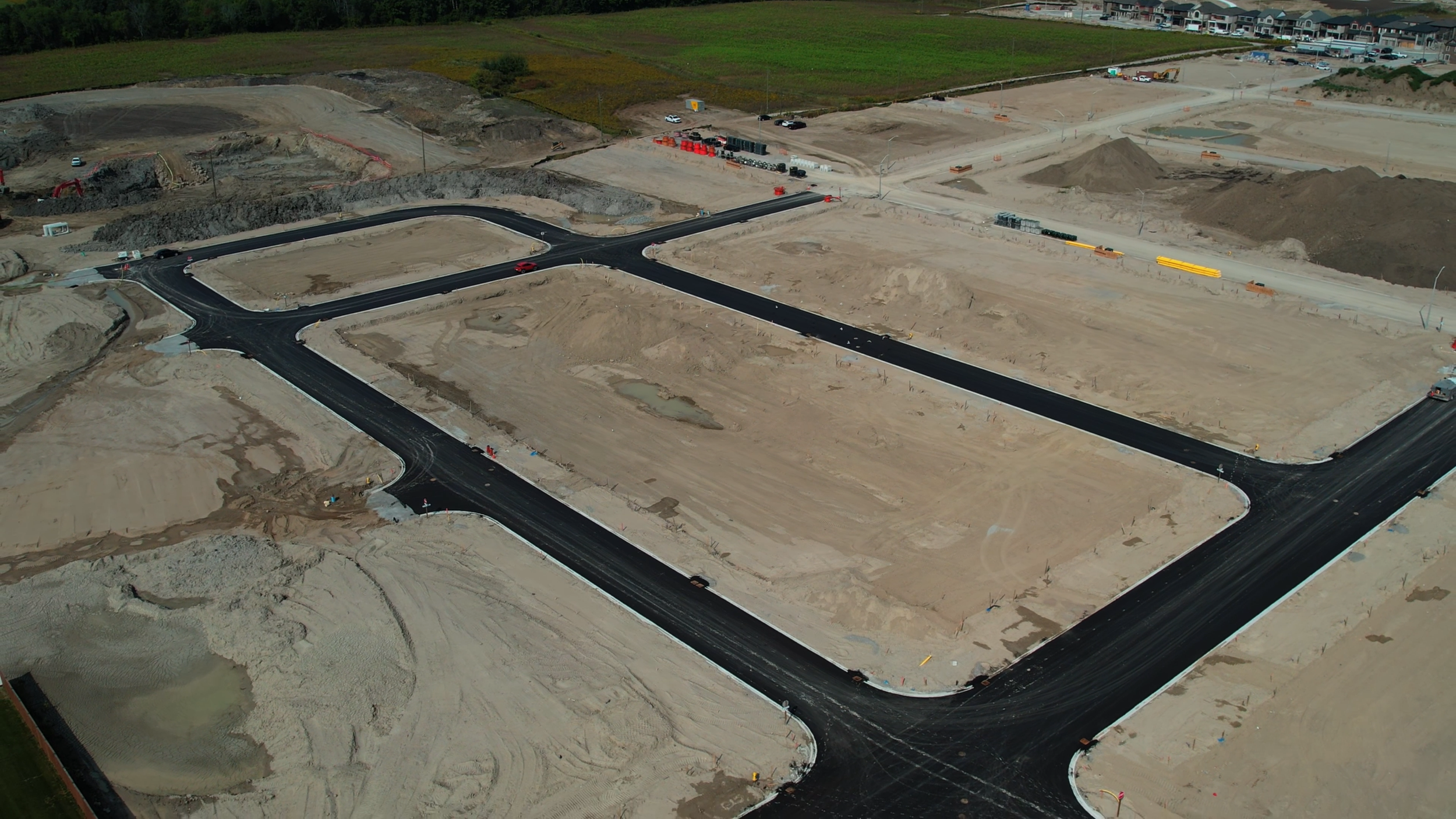 An aerial view of a construction site with newly paved roads, dirt mounds, and some vehicles, surrounded by fields and a residential area in the background.