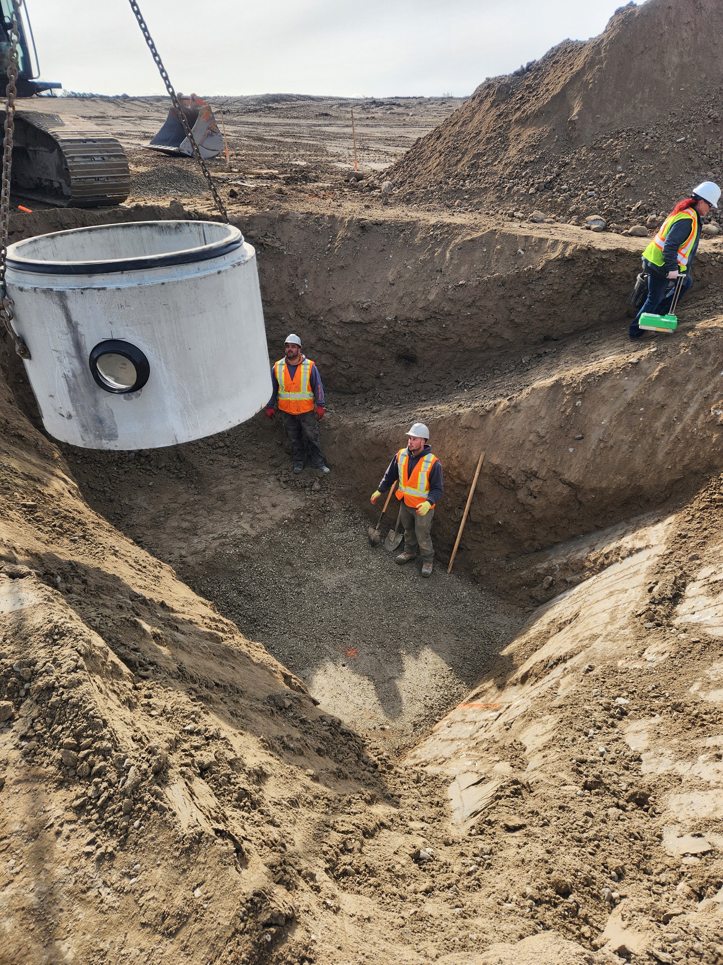 Construction workers in safety vests and helmets installing a large concrete pipe in a dug trench at an outdoor construction site.