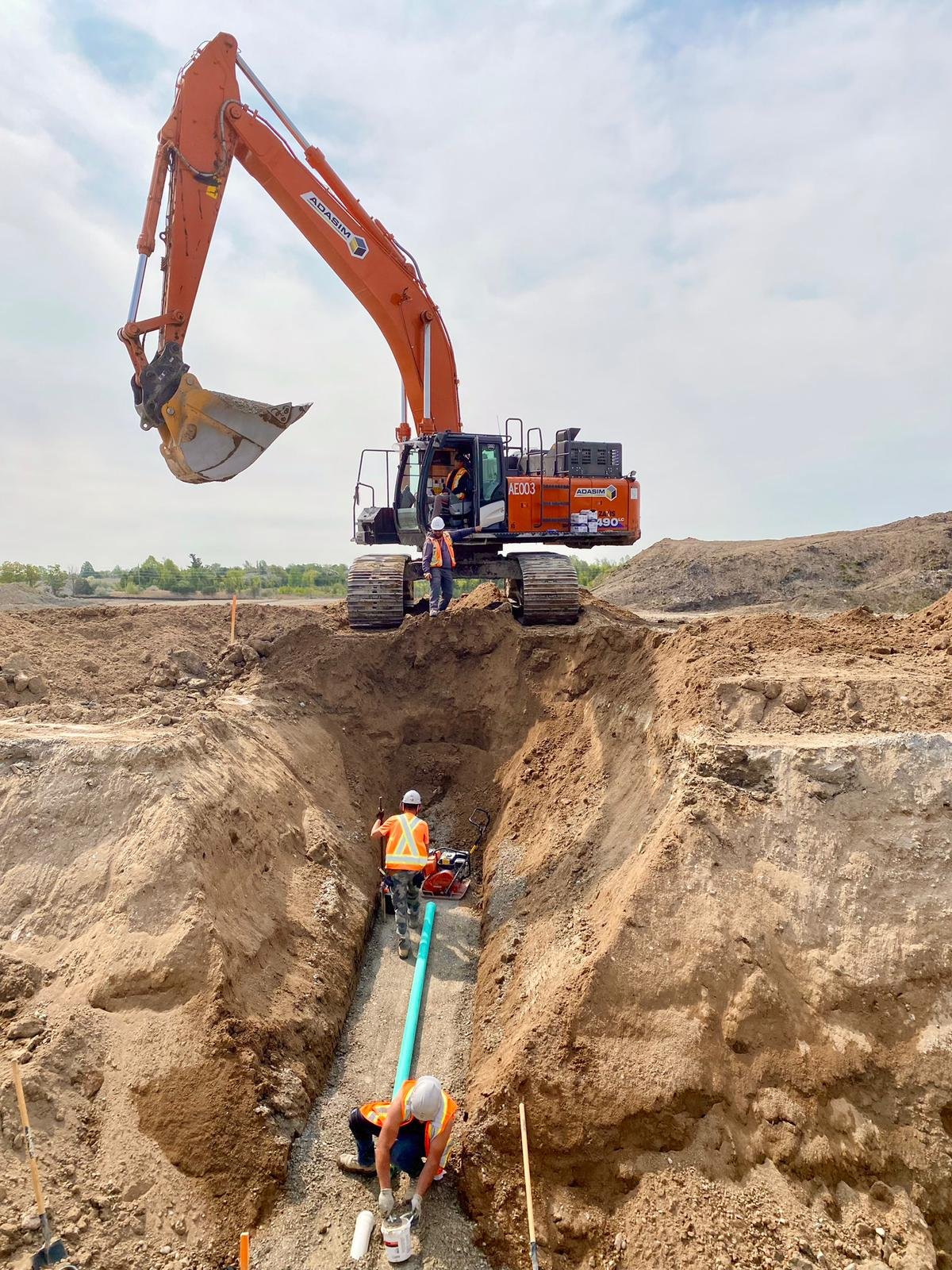 Construction workers install an underground pipe using a large orange excavator on a construction site.