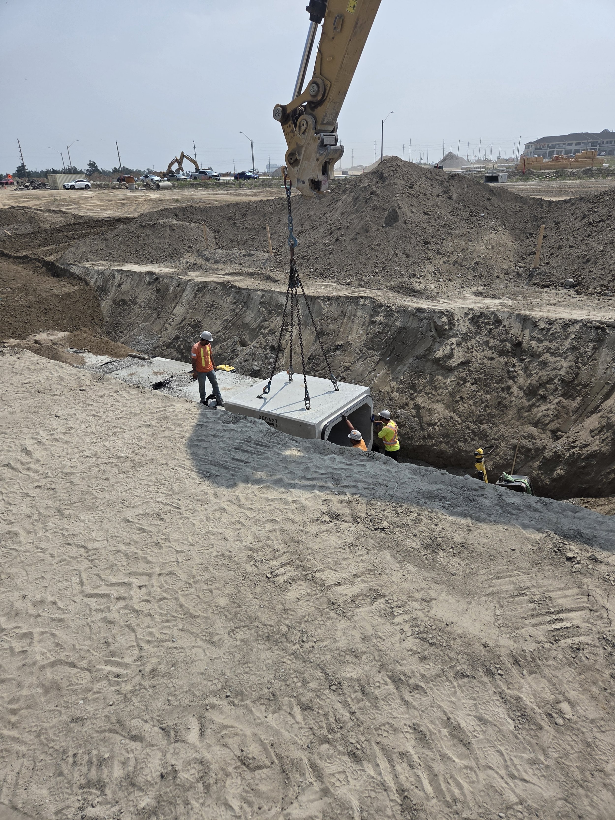 Workers at a construction site lift a large concrete slab with an excavator, preparing to place it into a trench.