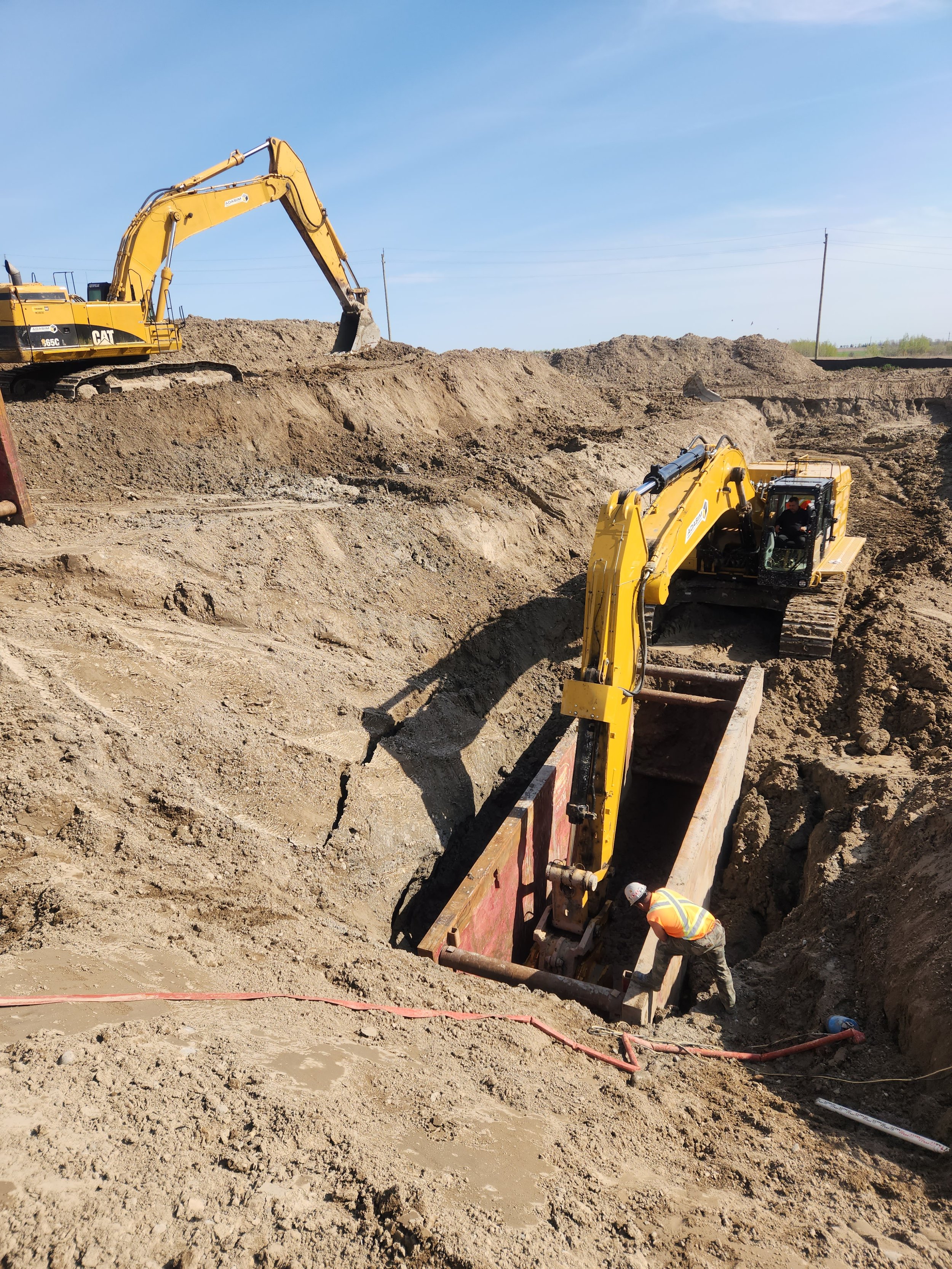 Two yellow excavators digging a trench, with a construction worker in an orange safety vest working at the bottom of the trench, under a clear blue sky.