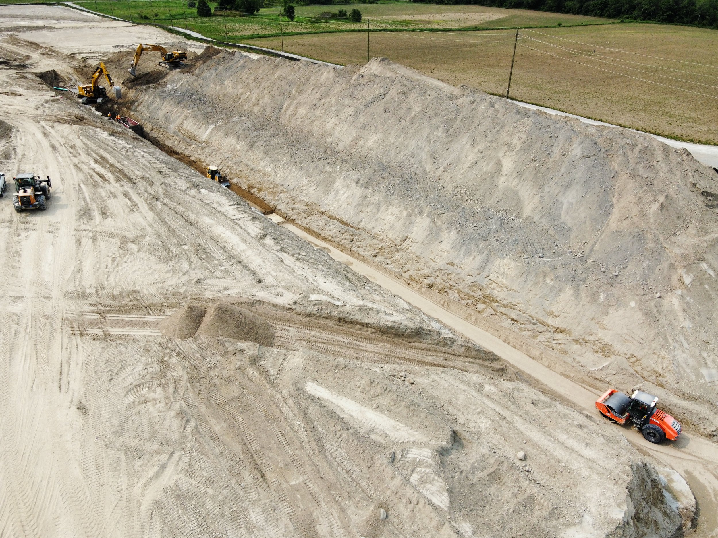 Construction site with multiple excavators working on earthwork, large dirt slope with graders, and trucks moving soil; green fields in the background.