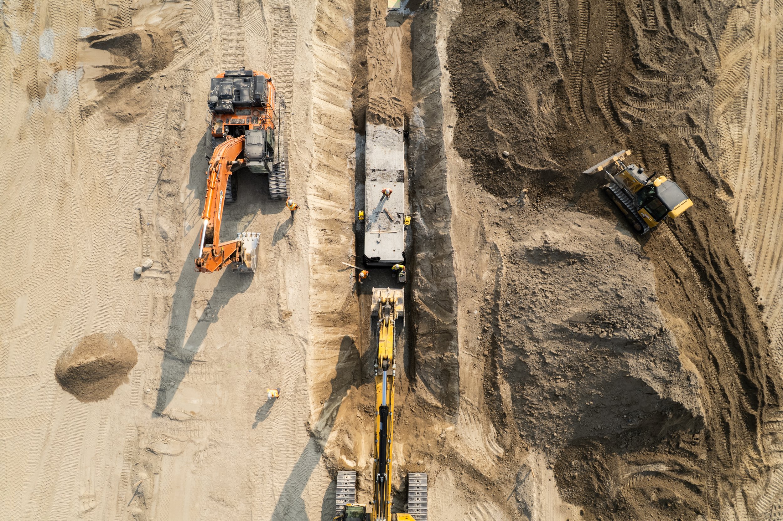 Aerial view of a construction site with excavators and workers working on a trench for laying pipes or cables, surrounded by mounds of dirt and construction equipment.