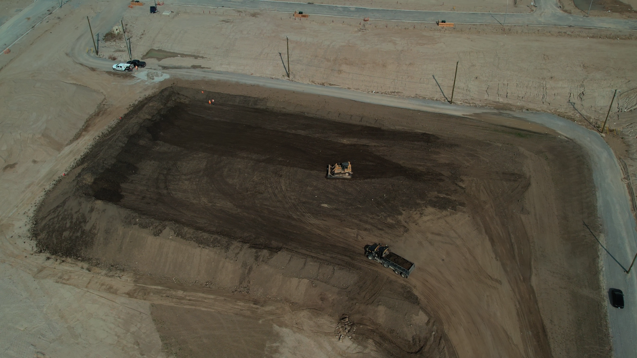 An aerial view of a construction site with dirt being leveled by construction vehicles, surrounded by partially paved roads and utility poles.