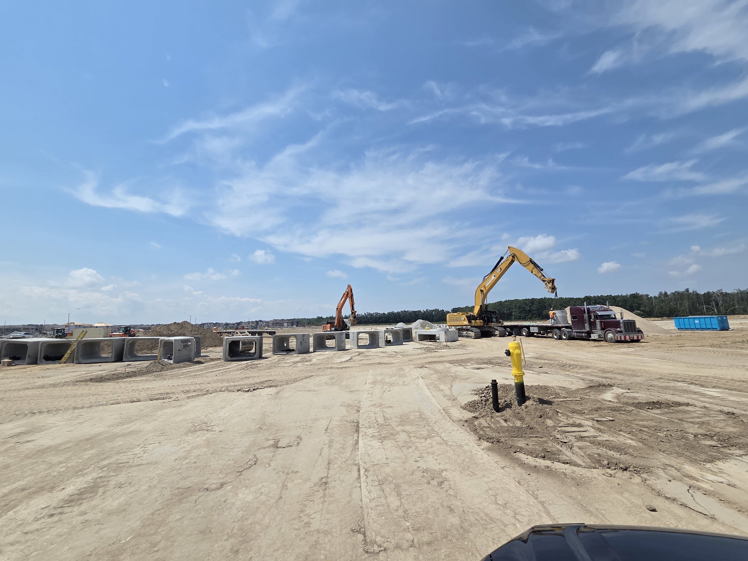Construction site with heavy machinery, including cranes and trucks, on a dirt road under a partly cloudy sky.