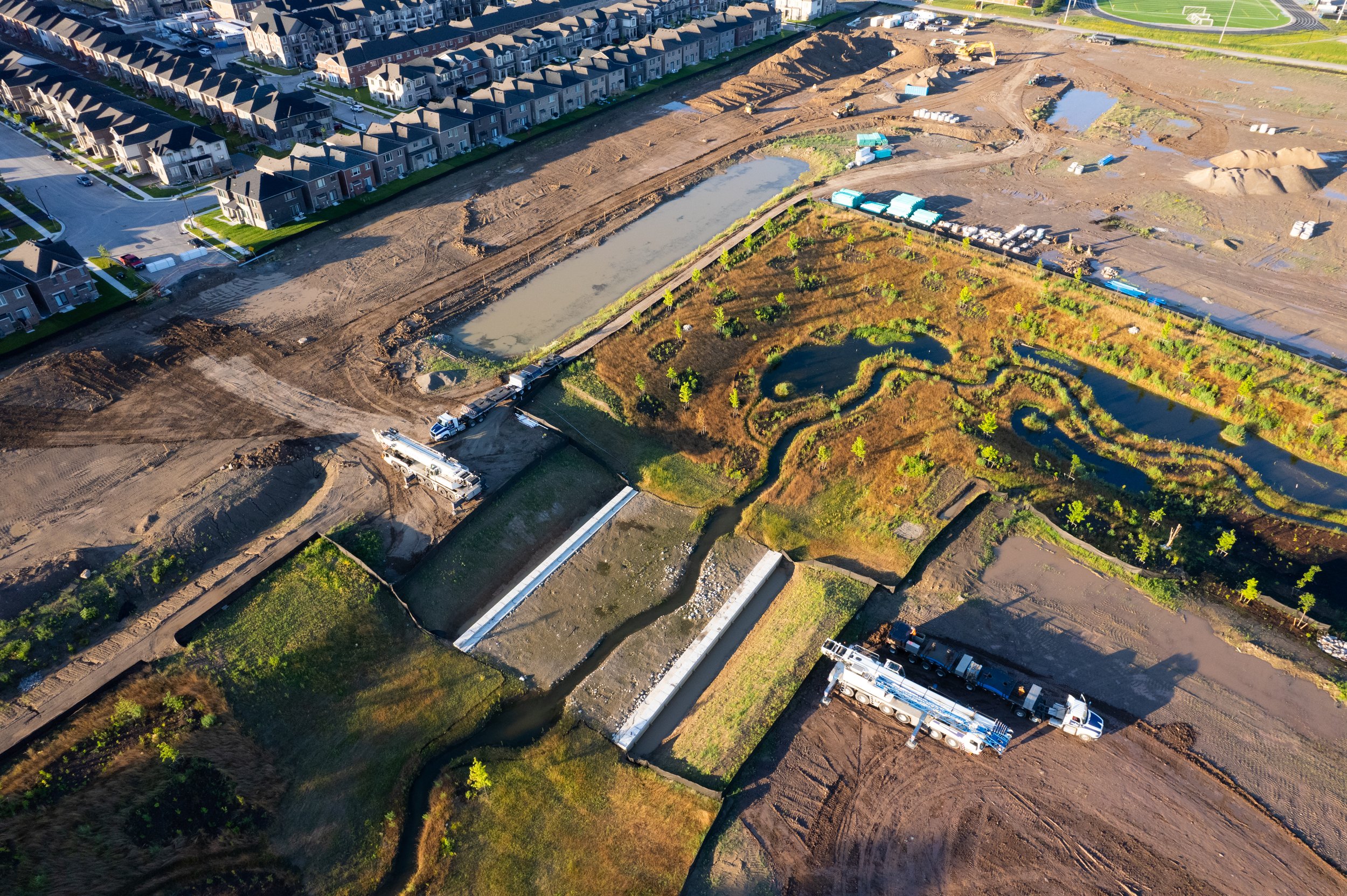 Aerial view of a construction site adjacent to a neighborhood with multiple newly built houses, showing ongoing work, water bodies, and vehicle trucks.