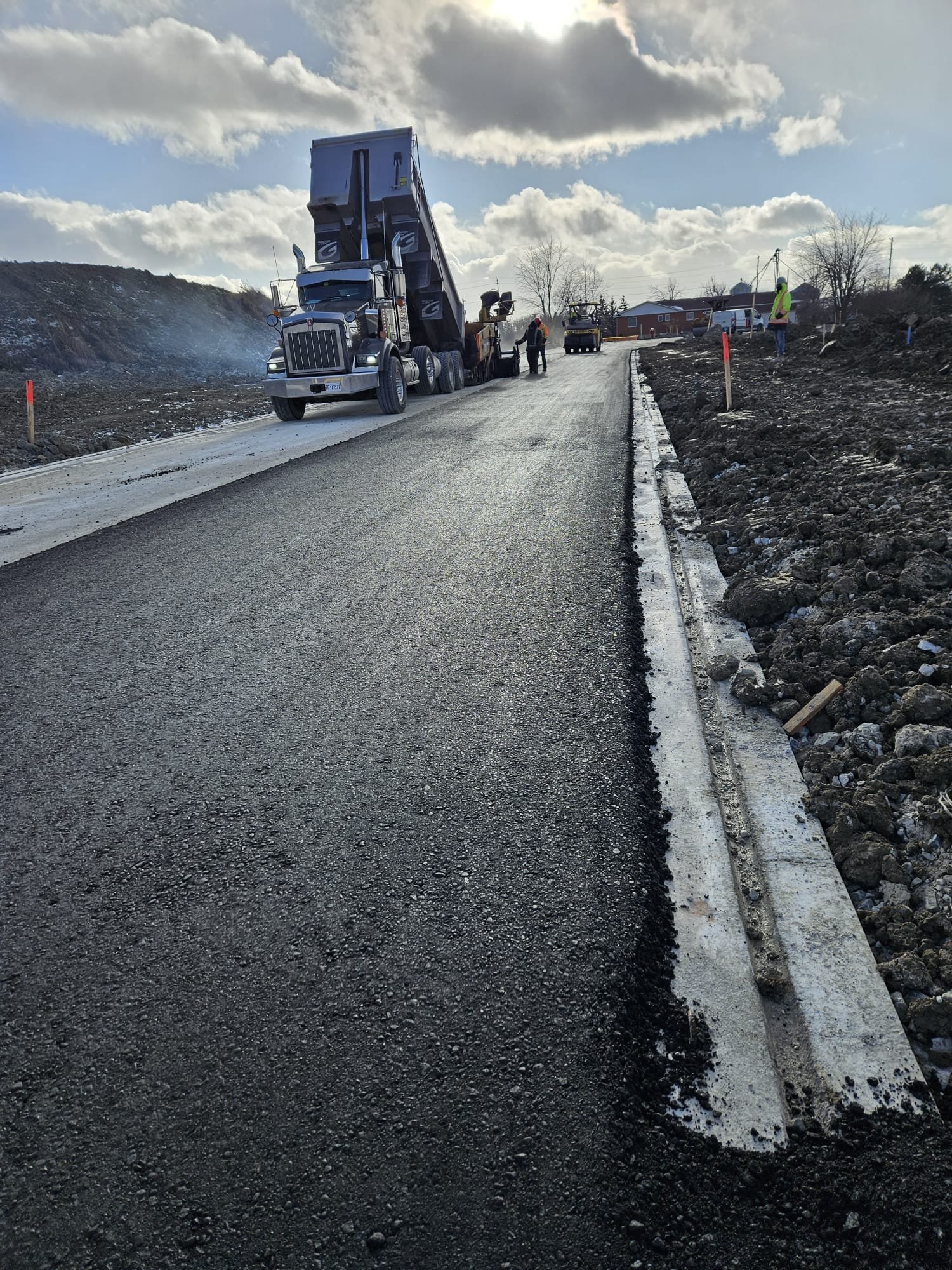 Construction workers paving a road with asphalt, with a large truck and construction vehicles in the background under a partly cloudy sky.