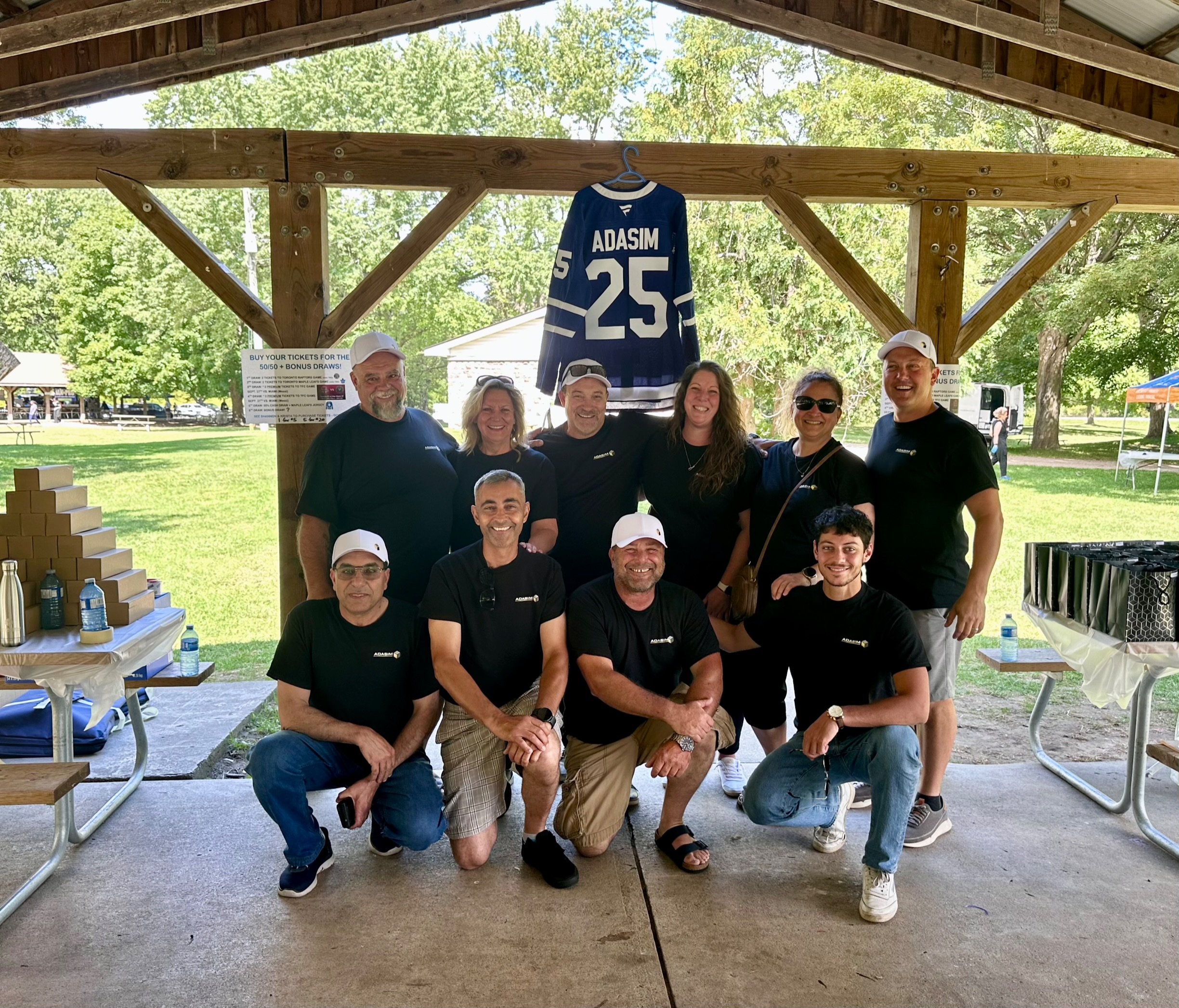 Group of ten people posing under a wooden pavilion with a hockey jersey hanging behind them. The group includes five men and five women, smiling for the photo. The setting appears to be outdoors in a park or fairground with trees and tents visible in