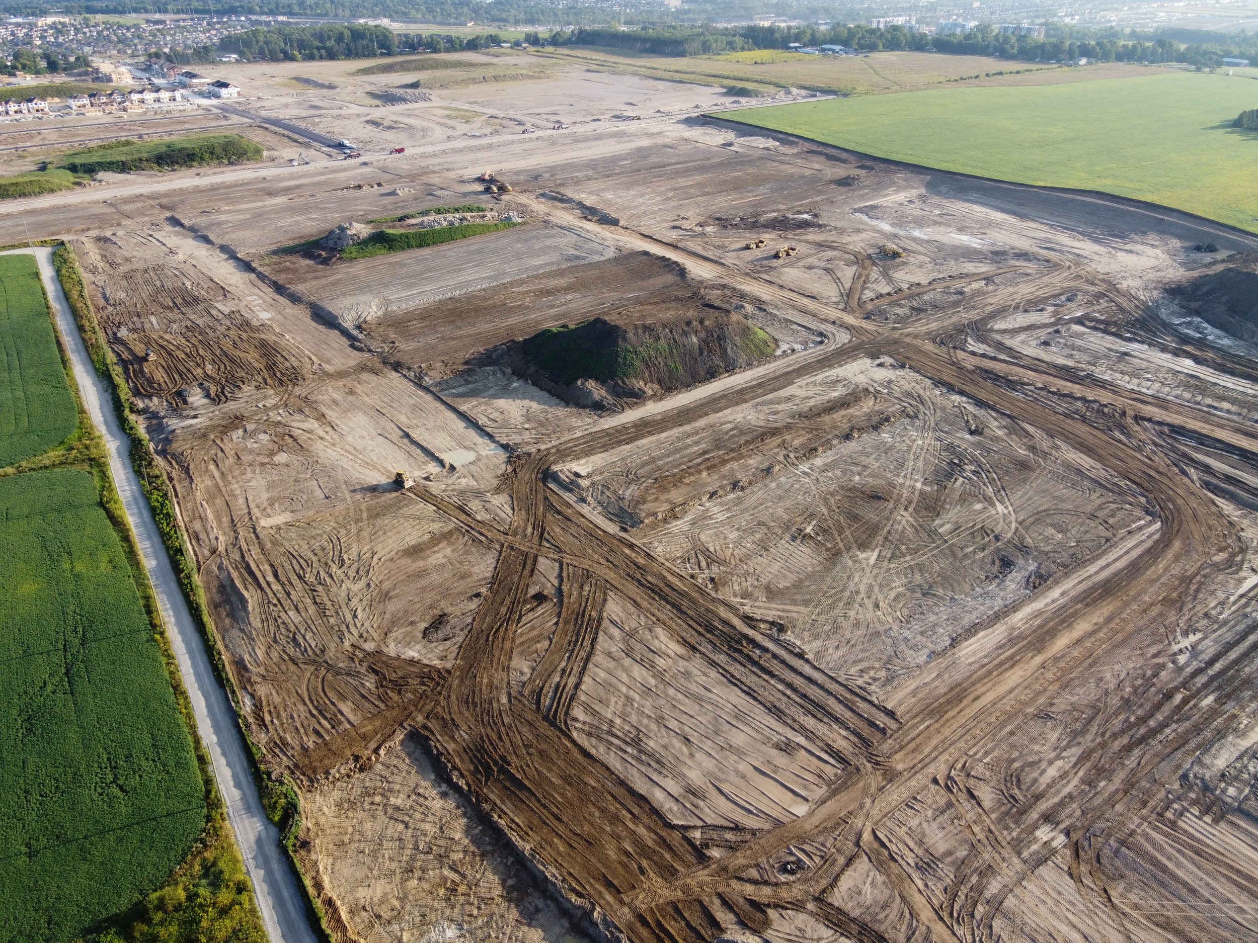 Aerial view of a construction site with cleared and excavated land, surrounded by grassy fields and some residential buildings in the background.