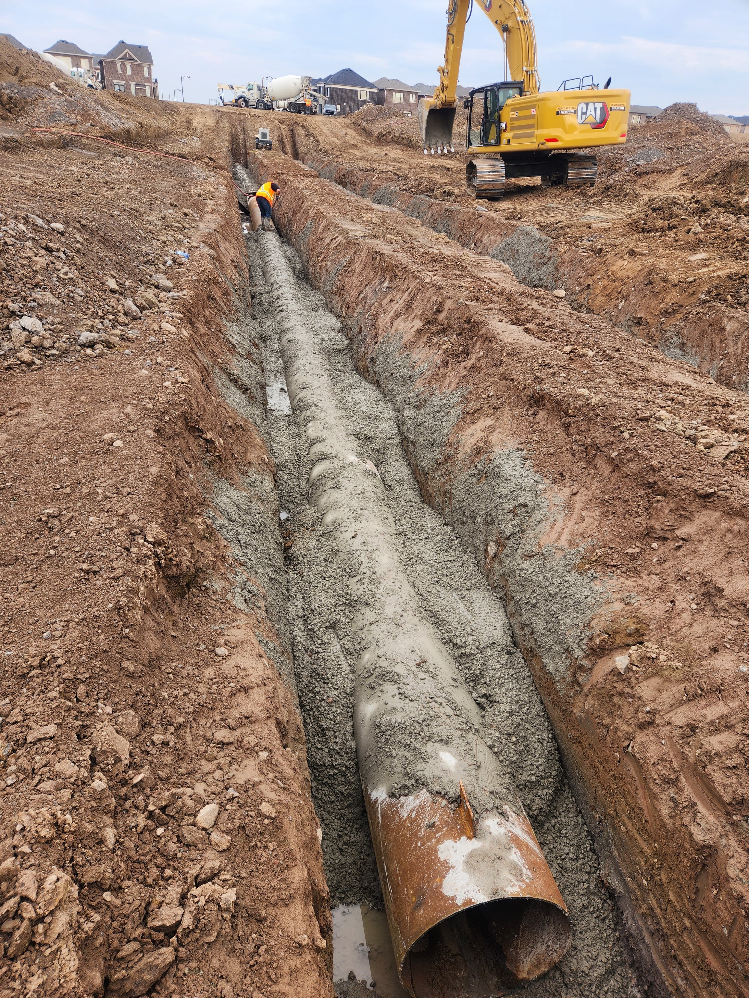 Construction site with an excavator and worker installing a large pipe in a trench during daylight.