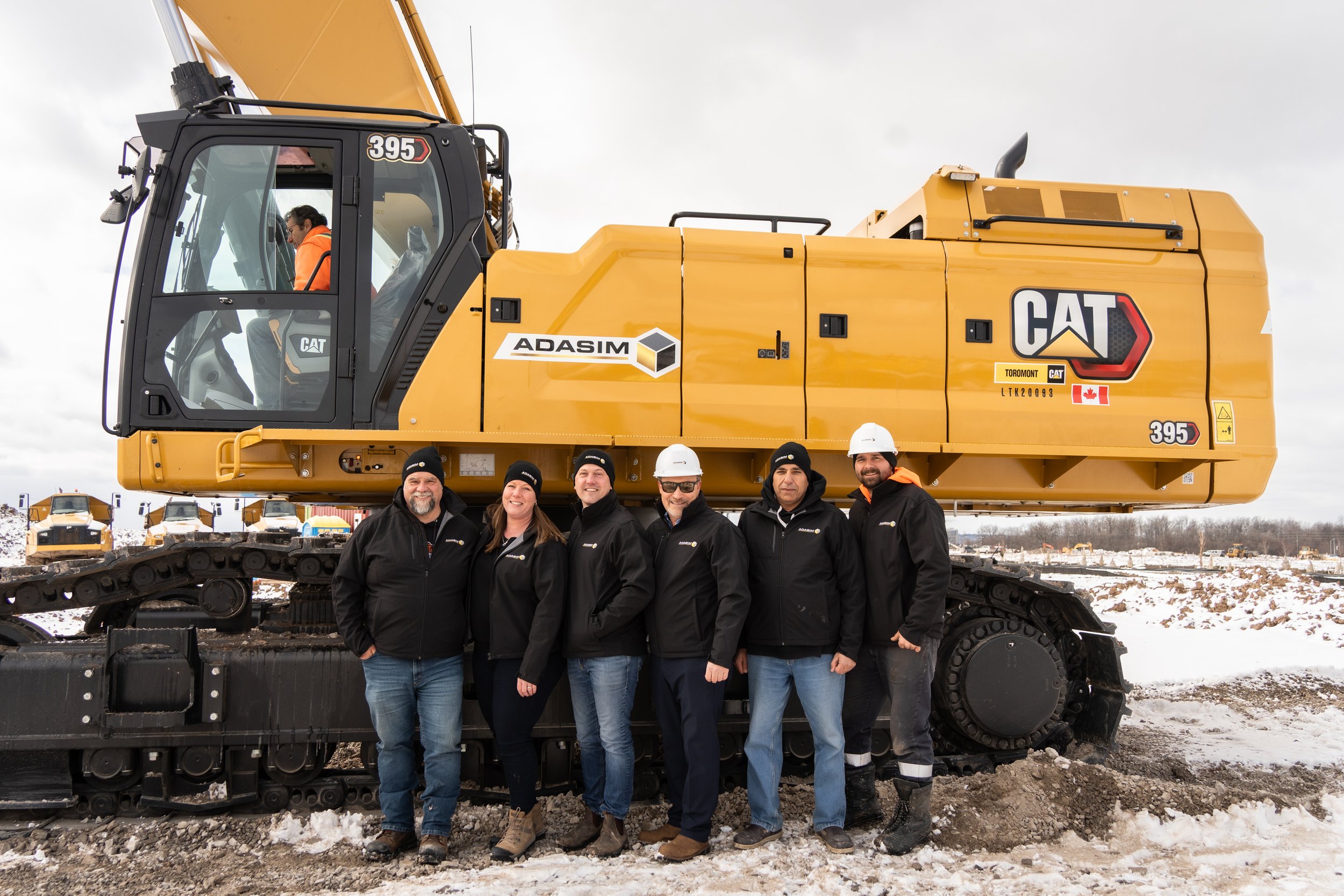 Six people standing in front of a large yellow CAT construction excavator outdoors in a snowy area. They are wearing black jackets and hats, with some in white hard hats. The excavator has various logos and labels, including ADASIM and a Canadian fla