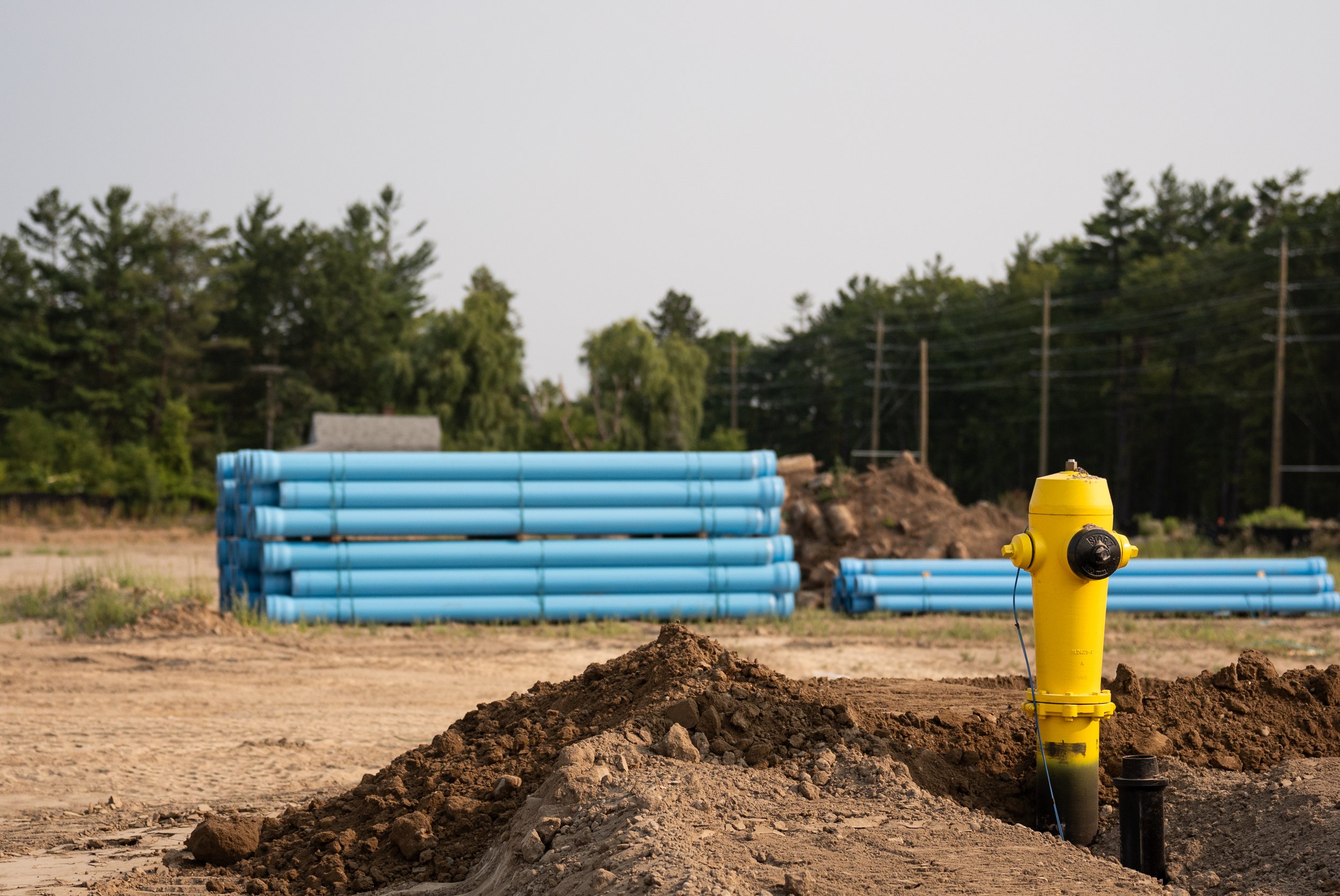 Construction site with a yellow fire hydrant in the foreground, dirt piles, and blue pipes stacked in the background, with trees and power lines behind.
