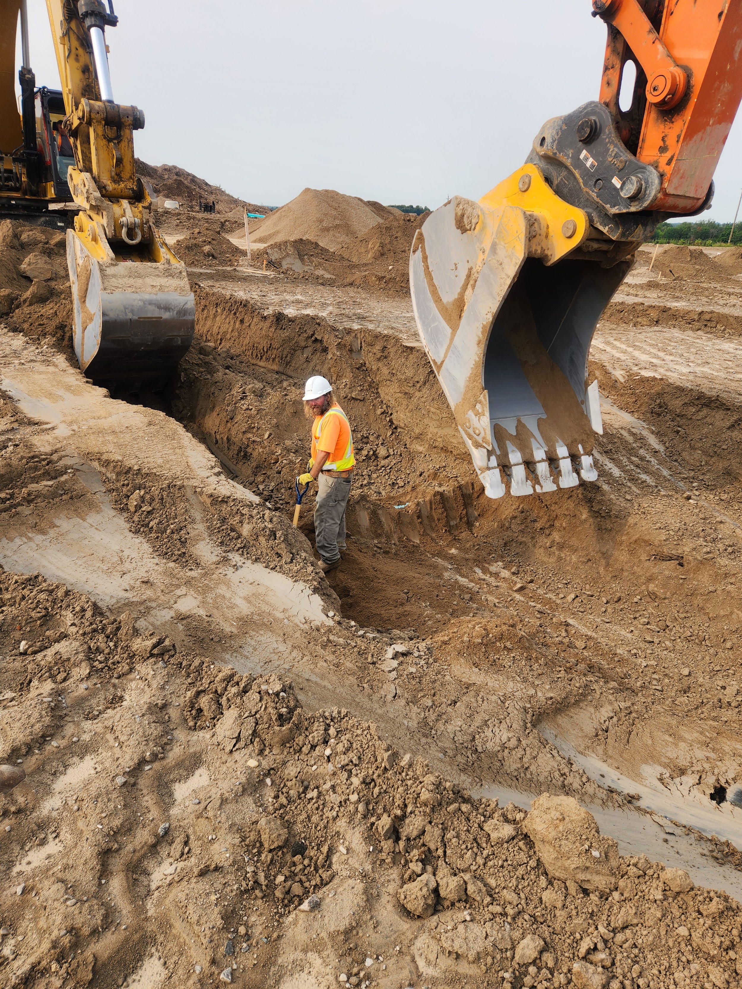 A construction worker in a white hard hat and orange shirt working in a trench with large excavator buckets overhead at a construction site.