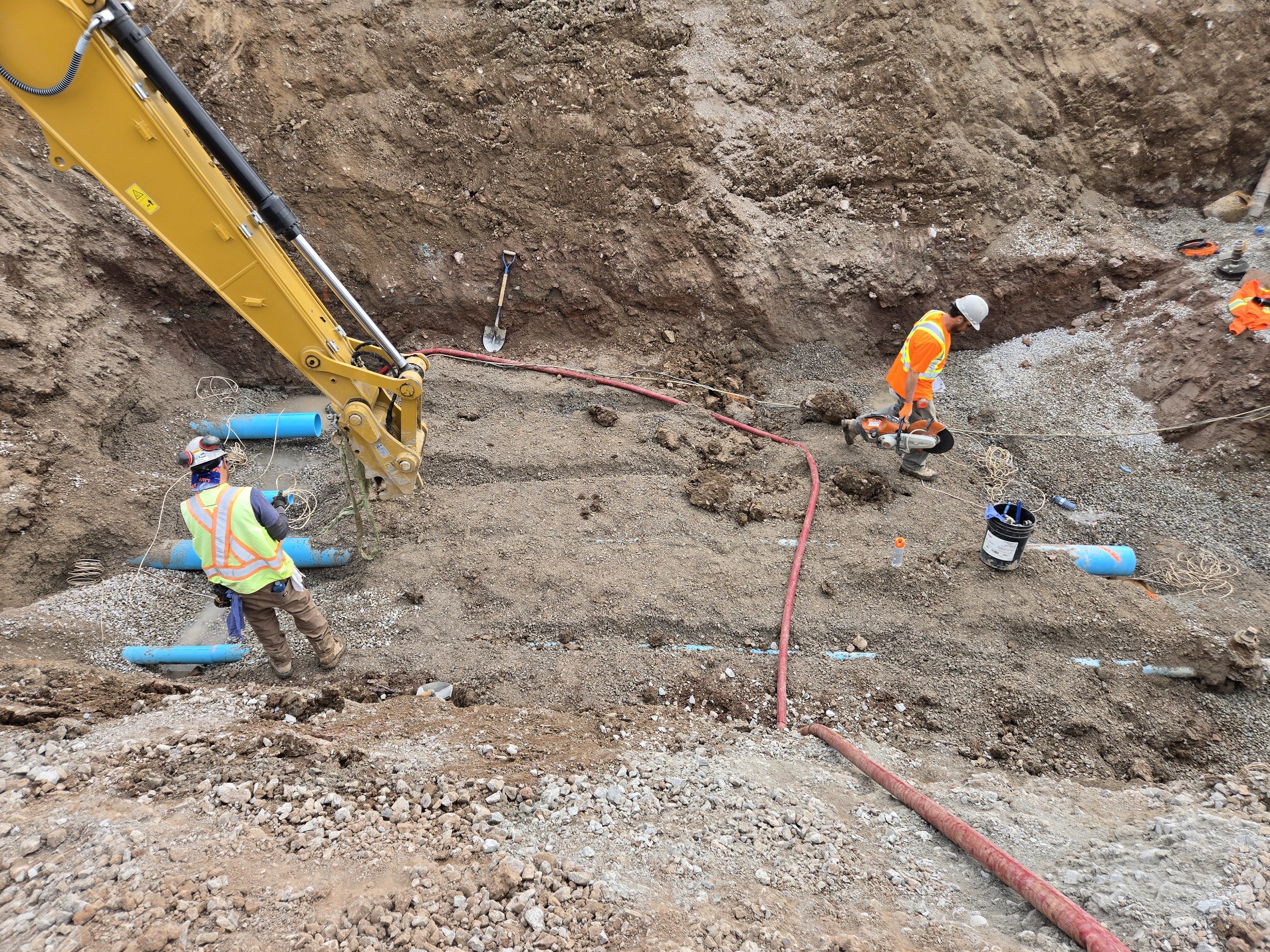 Construction workers installing blue pipes in an excavation site, with one worker using a handheld power tool, and a yellow excavator arm nearby.
