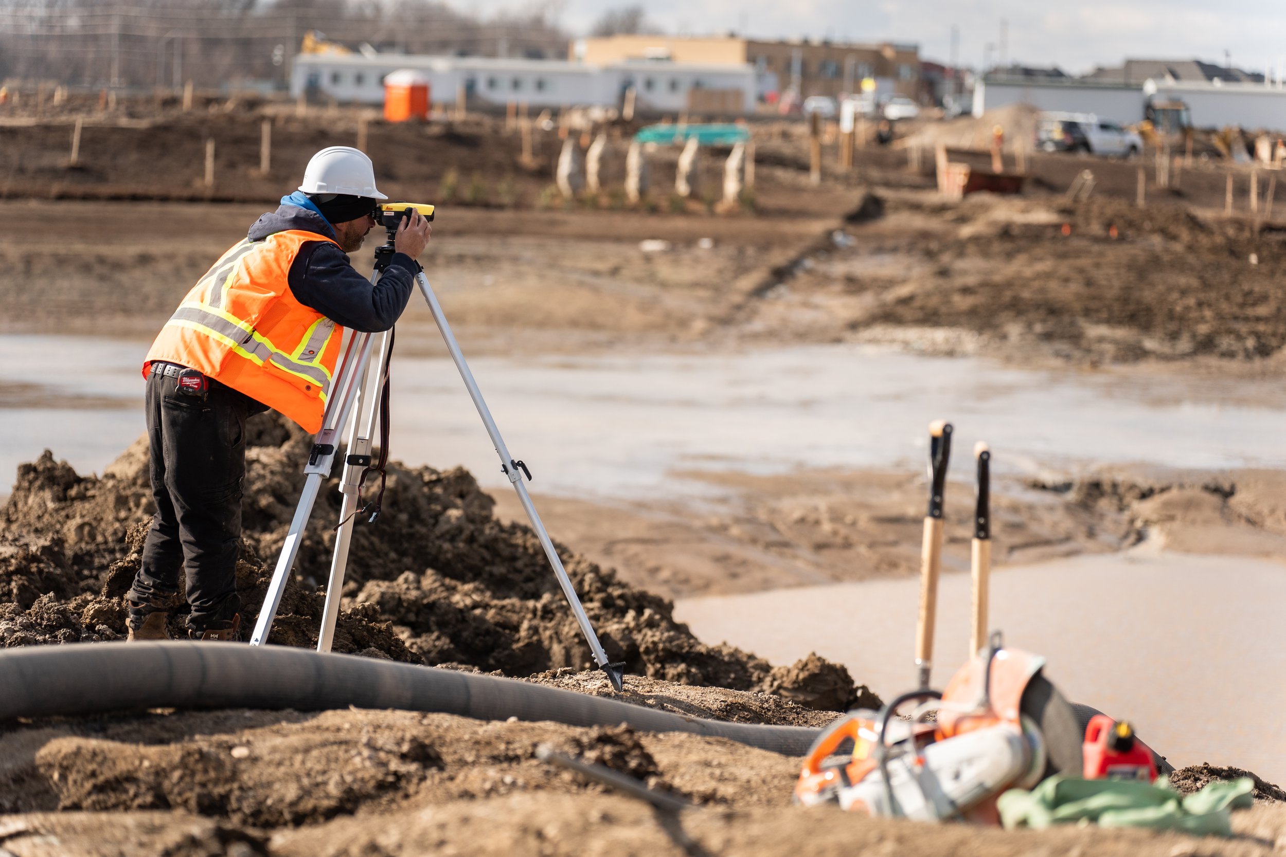 Construction worker using a surveying instrument on a construction site with building materials and equipment in the background.