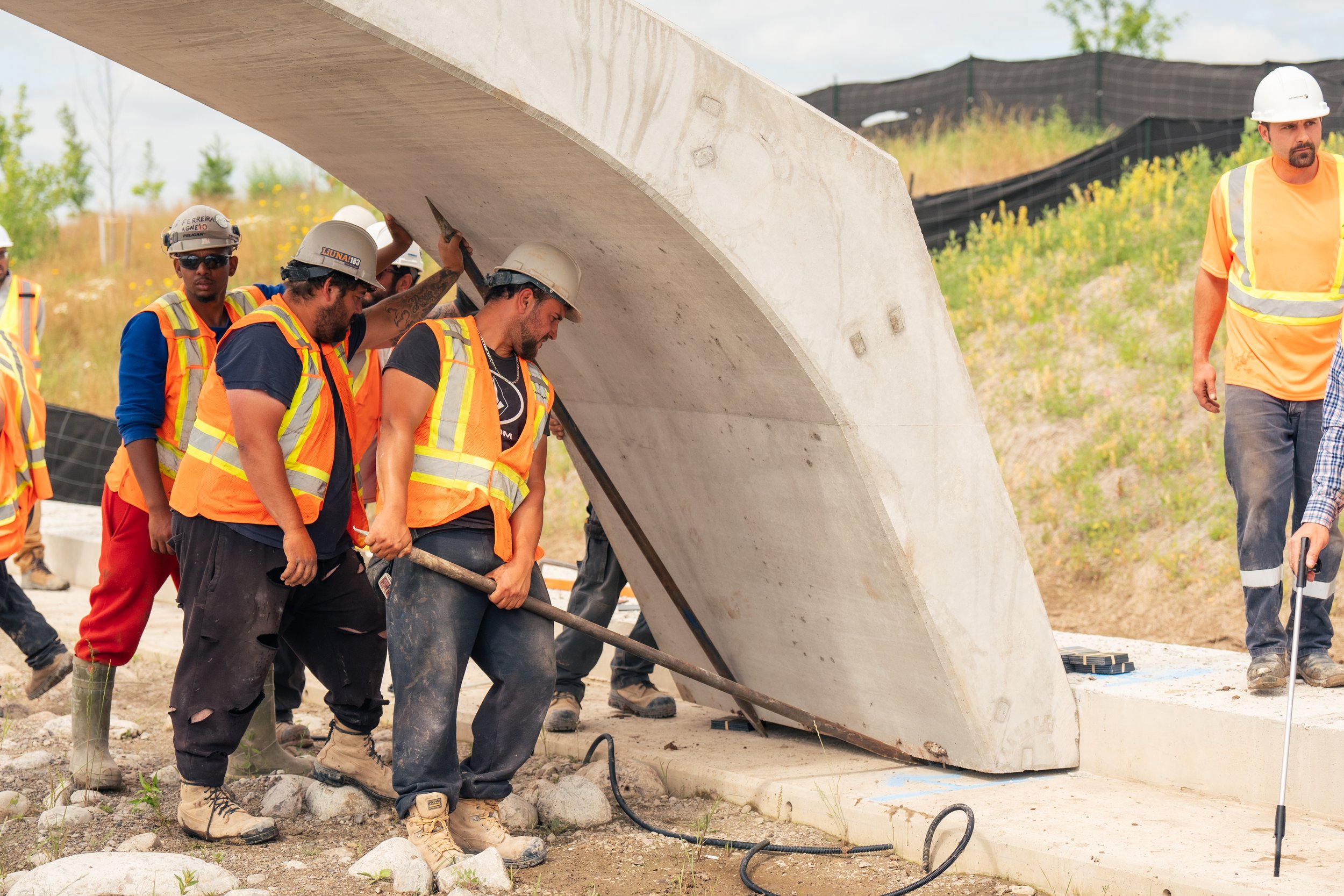 Construction workers in safety gear lifting and positioning a large concrete panel with a steel rod at a construction site.