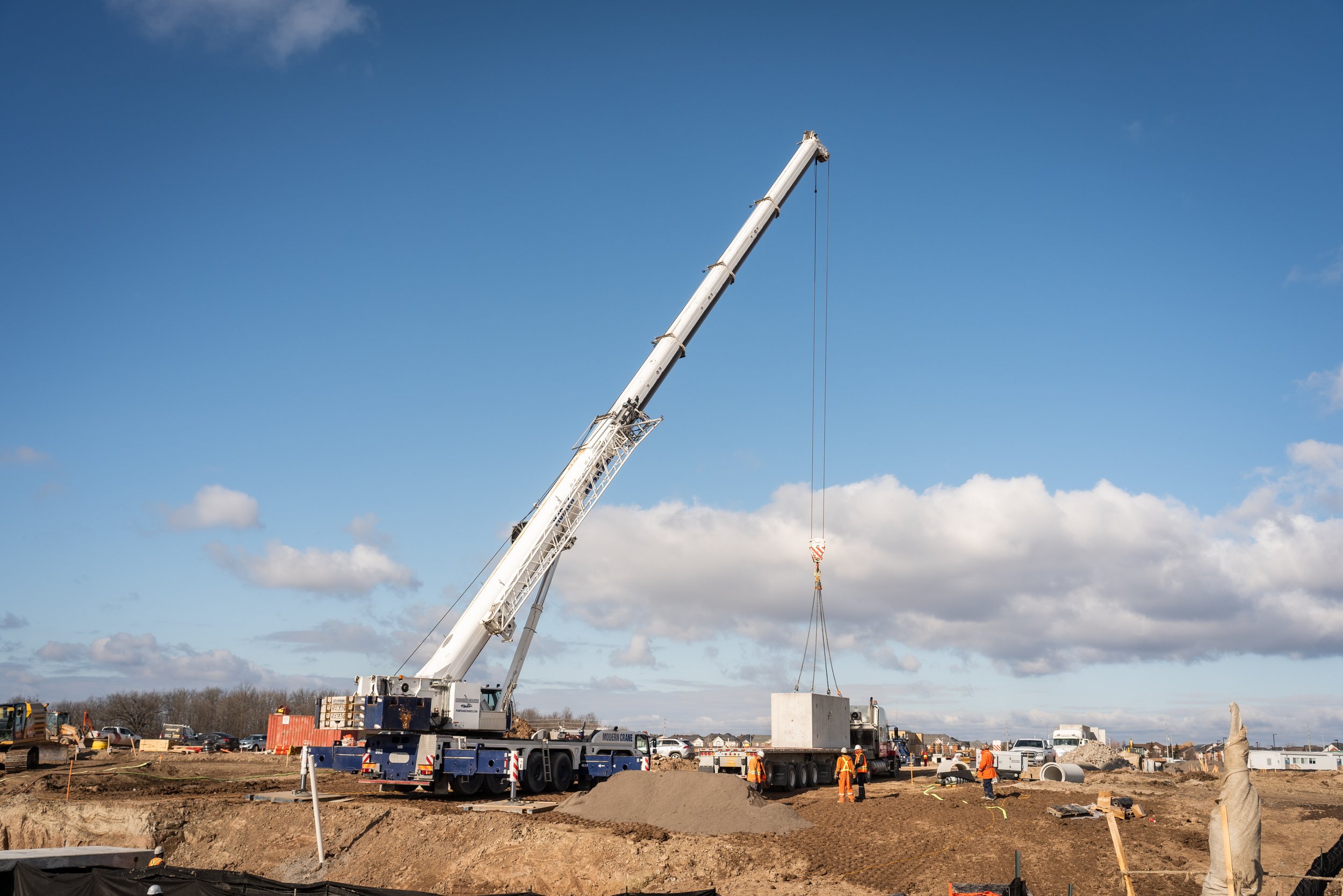 Construction site with a large crane lifting a concrete block, workers in orange safety vests and helmets, dirt and construction materials, with a blue sky and clouds overhead.