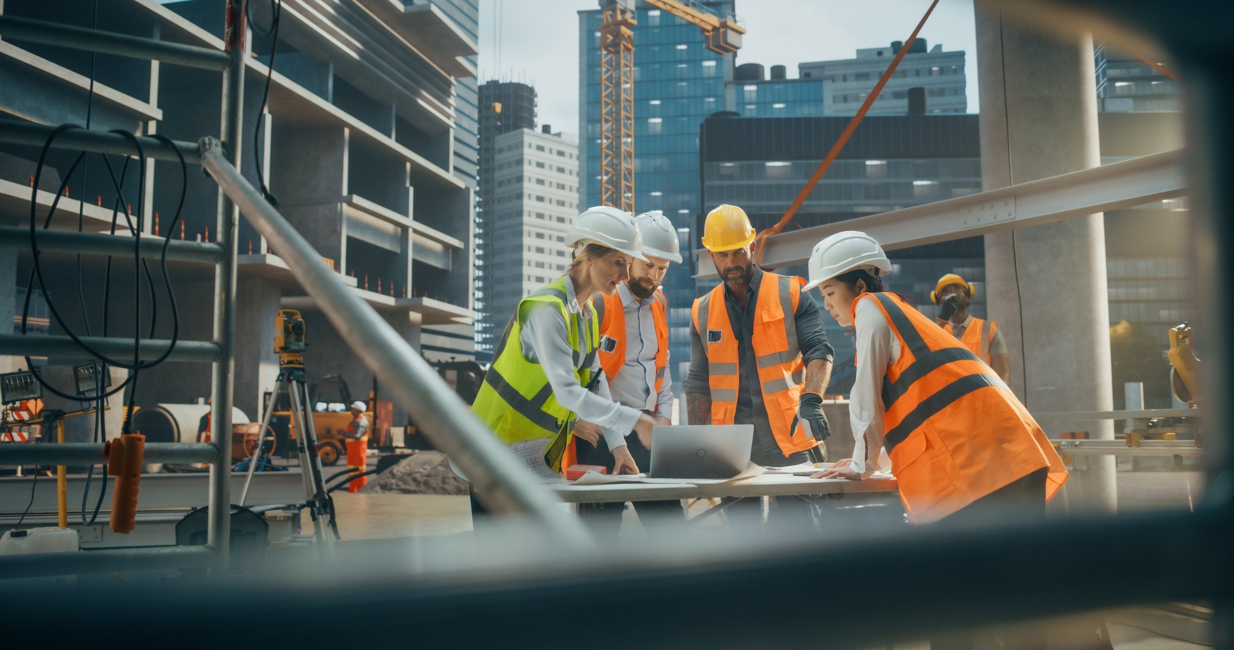 Construction workers in safety vests and helmets gathered around a table reviewing plans at a construction site with high-rise buildings and a crane in the background.