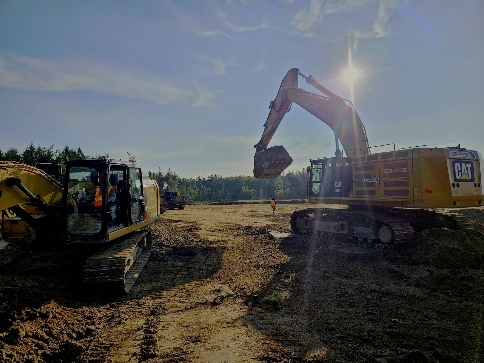 Construction site with two yellow Caterpillar excavators working on a dirt terrain, with workers and trucks in the background under a bright sky.
