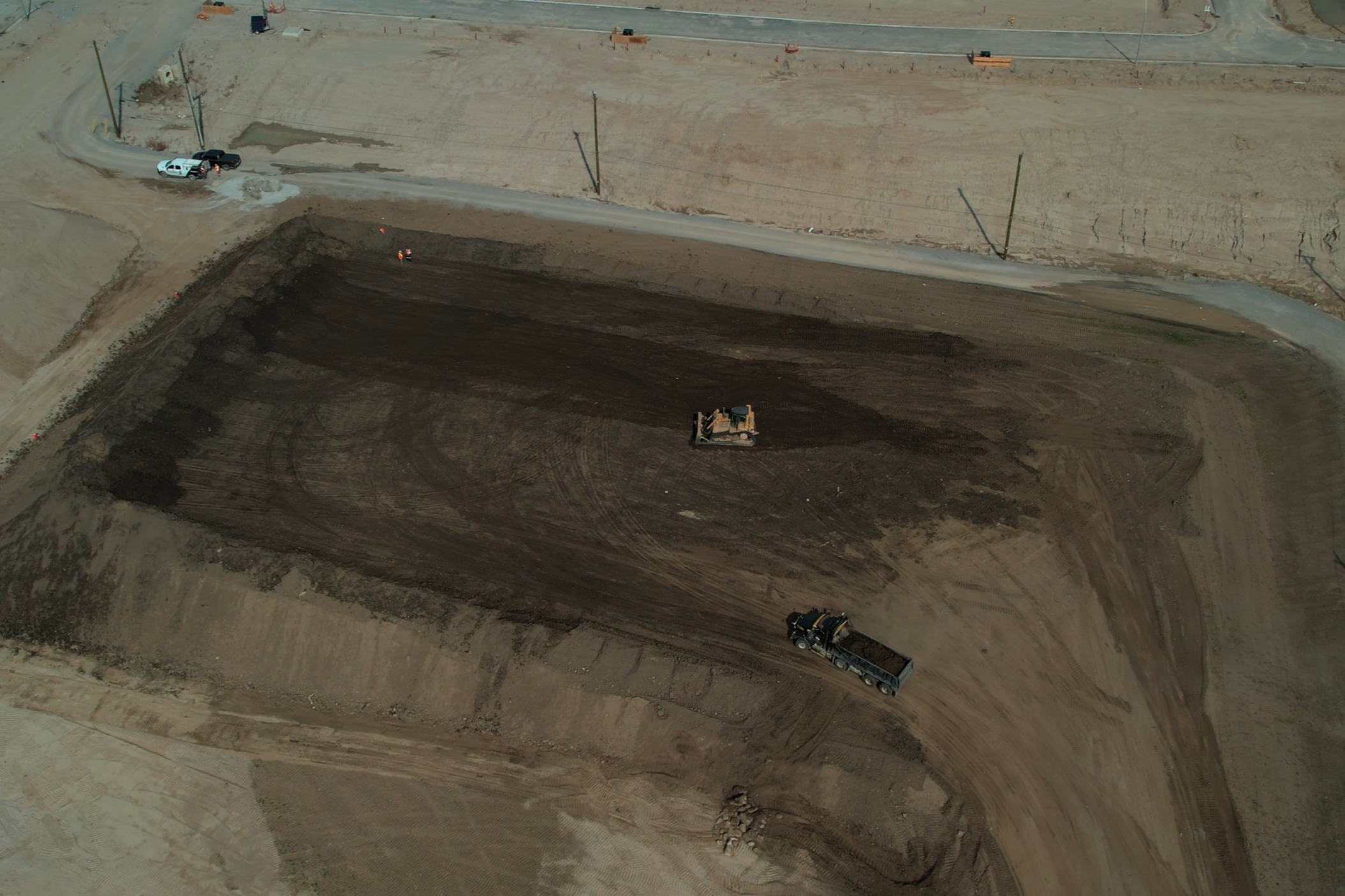 Construction site with excavator and dump truck on dirt area, and parked cars at the road edge.