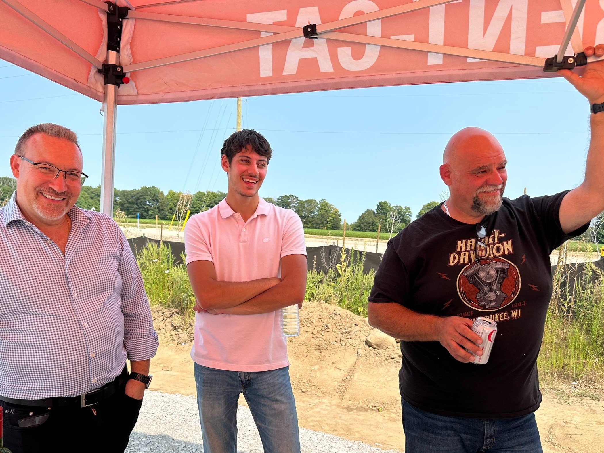 Three men standing under a pink canopy, smiling and talking outdoors with a blue sky, green fields, and a black fence in the background. The man on the right is holding a drink and adjusting the canopy. The man in the middle has arms crossed and is l