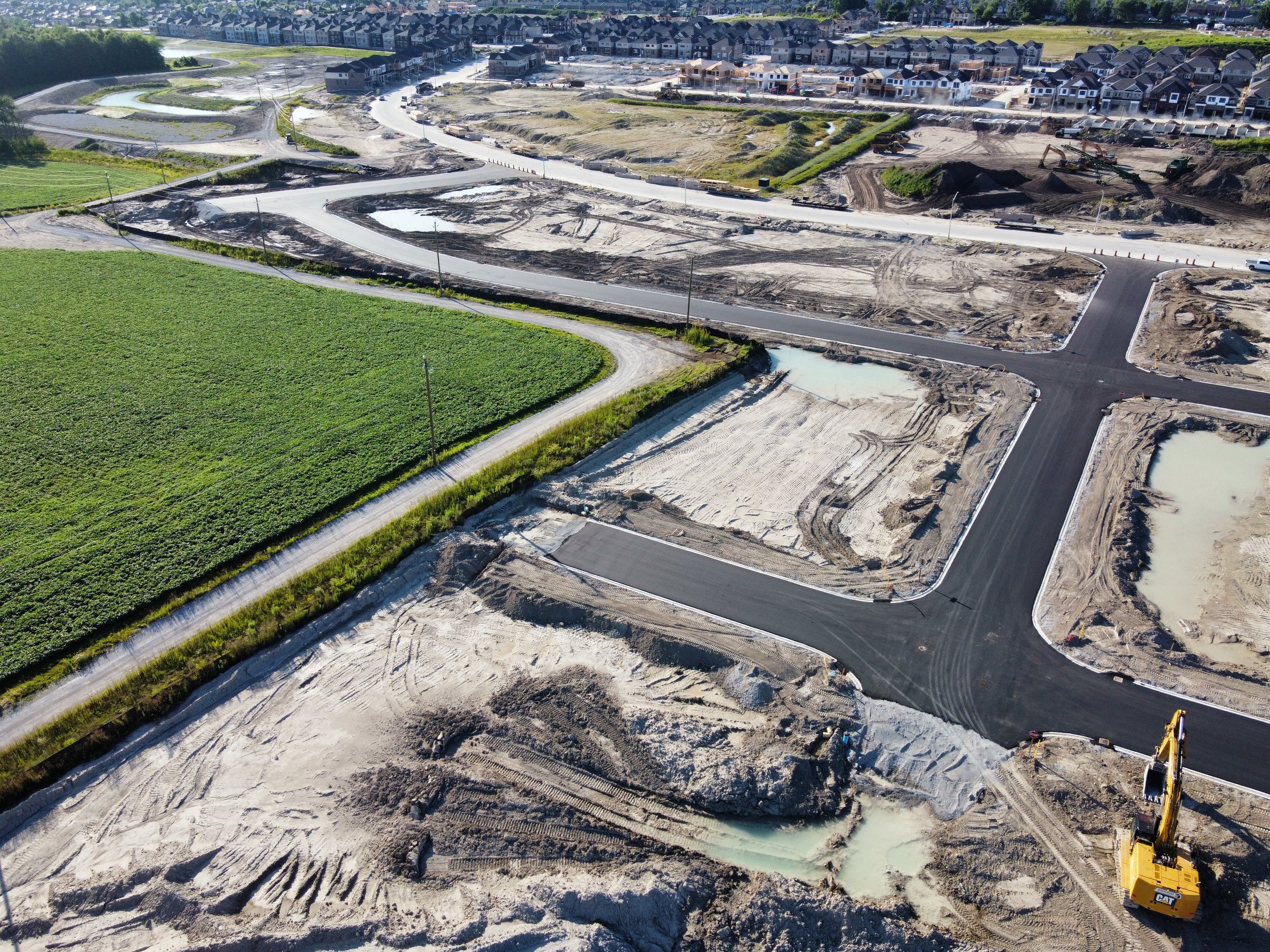 An aerial view of a construction site with newly paved roads, dirt, and construction equipment, including a yellow excavator, with residential houses in the background.